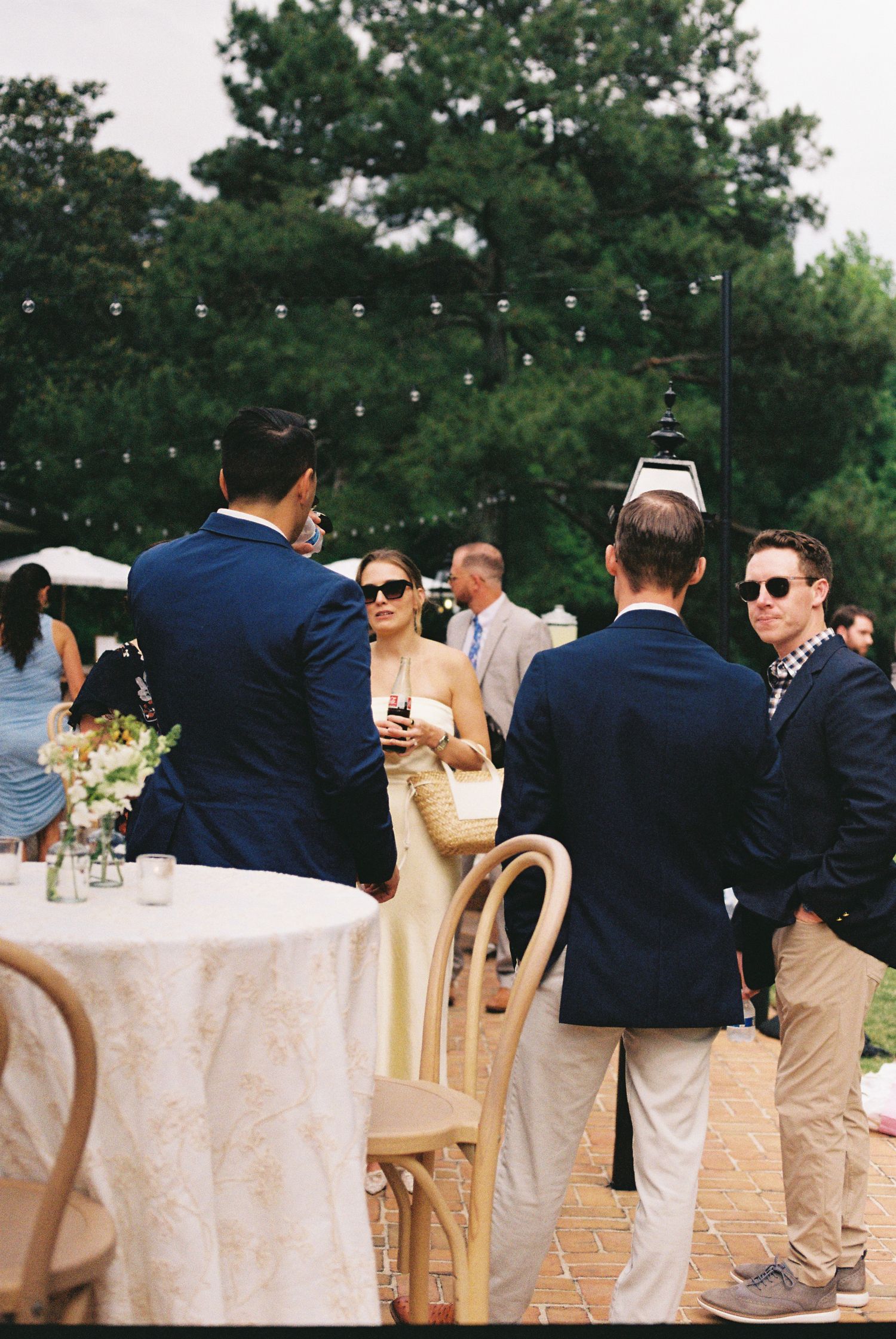 Guests in navy suits and summer dresses mingle at an outdoor wedding reception with string lights and lush greenery.