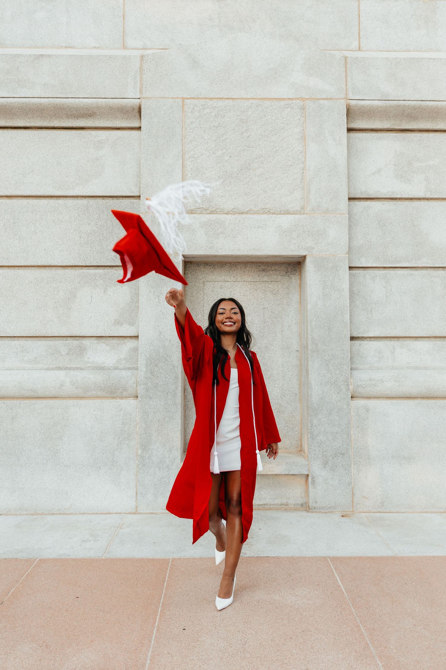 Smiling graduate in red cap and gown waving her cap in front of a stone building.