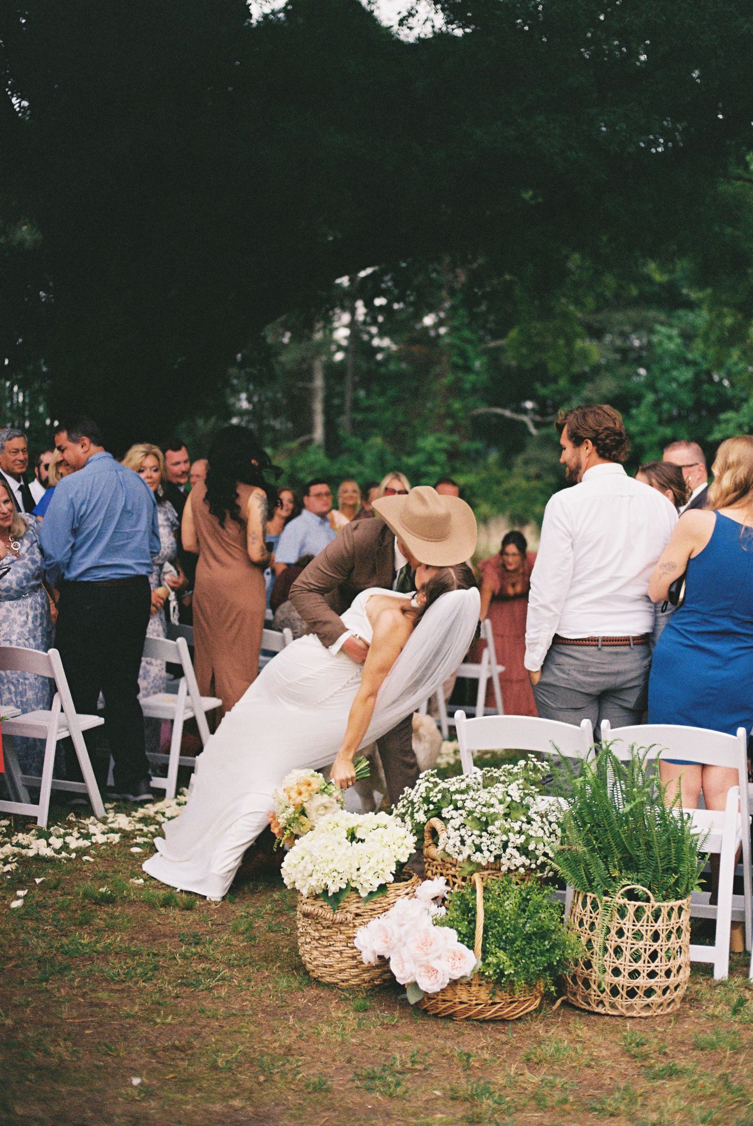 Bride and groom share a romantic dip kiss at outdoor wedding ceremony with floral basket arrangements.