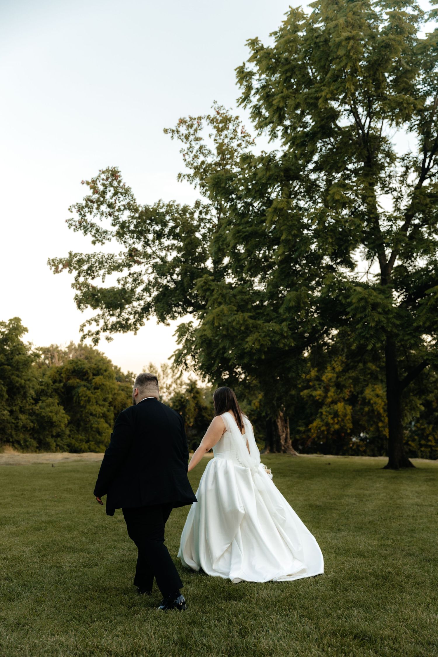 Couple walks hand in hand across grassy field during golden hour.