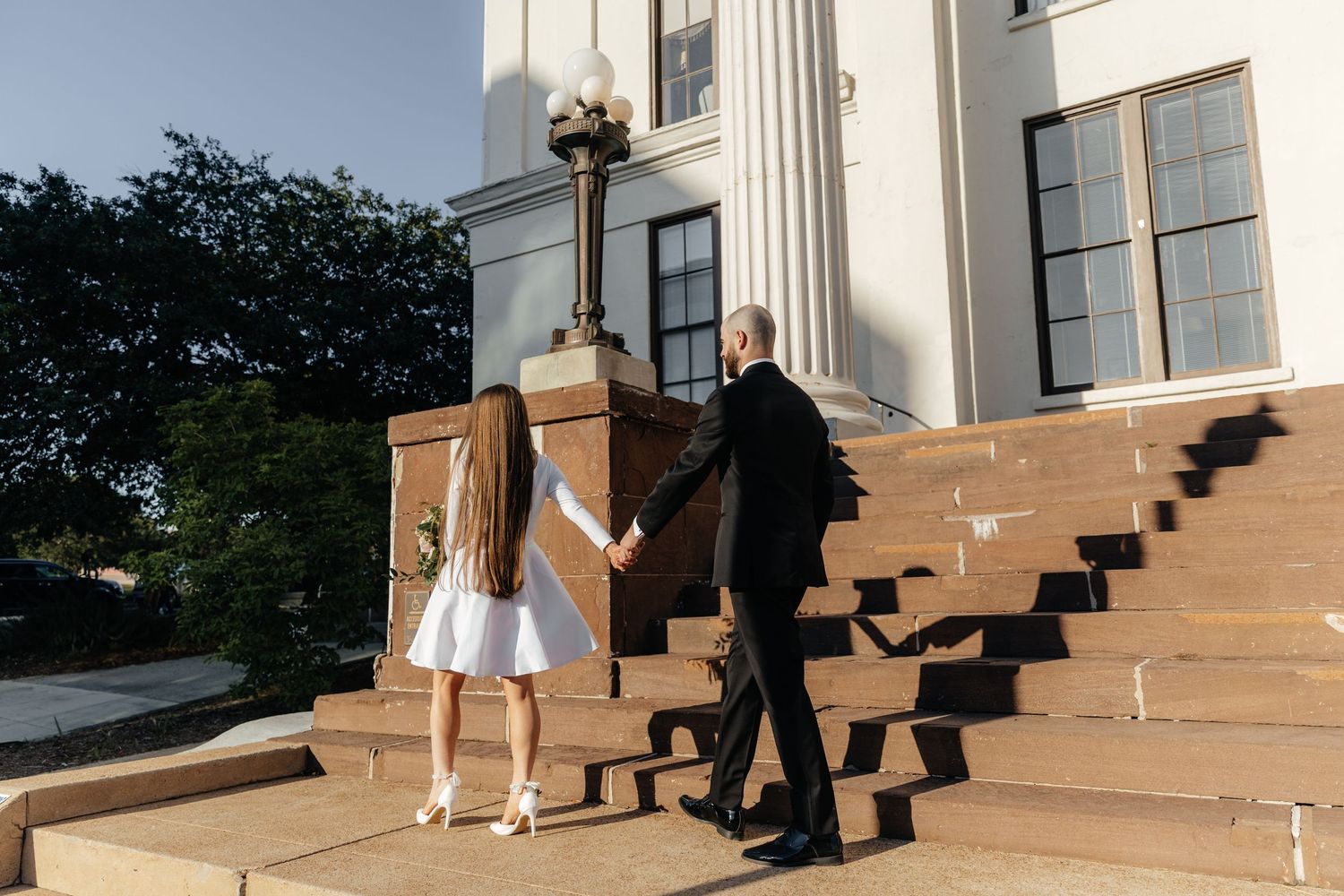 A couple in formal attire descends a grand staircase with white columns at a historic building.