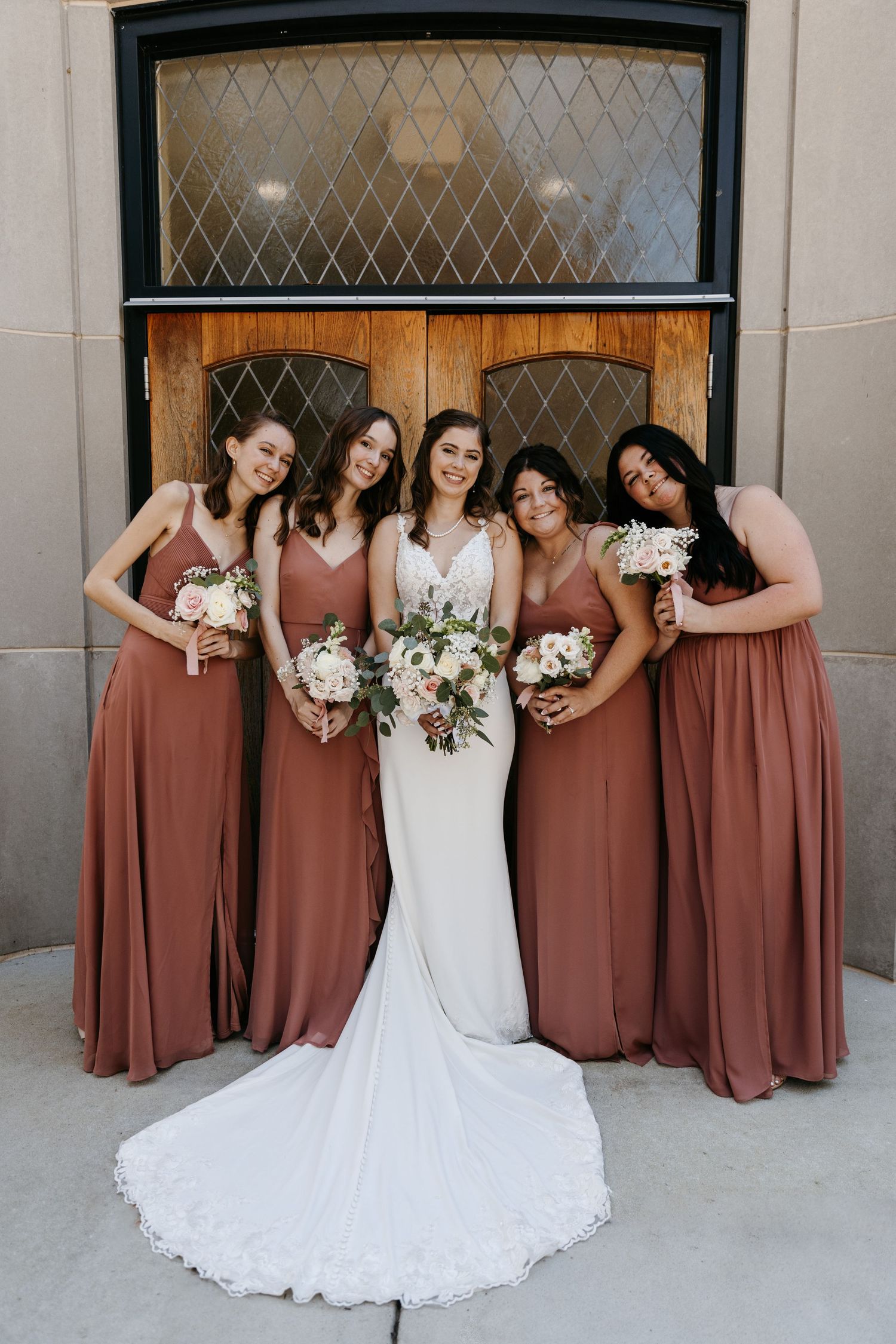 Bridal party poses playfully in front of brick building with bridesmaids in dusty rose dresses and bride in white gown.