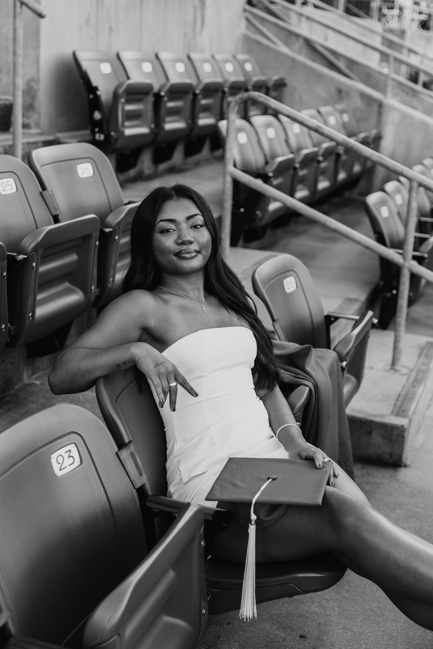 Black and white photo of a young woman in white dress holding graduation cap, seated in stadium bleachers.
