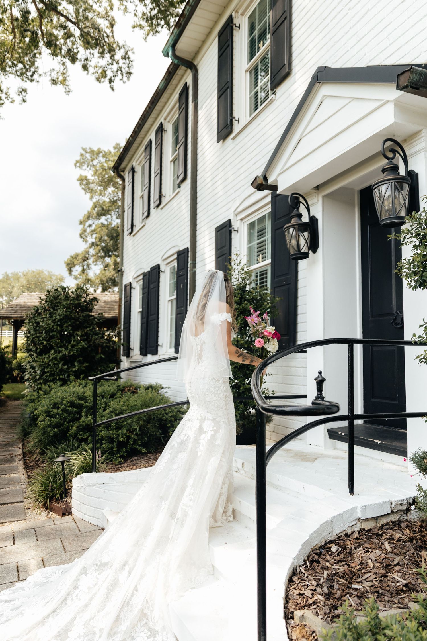 Bride in a lace mermaid gown and veil stands on steps of a white colonial home with black shutters.