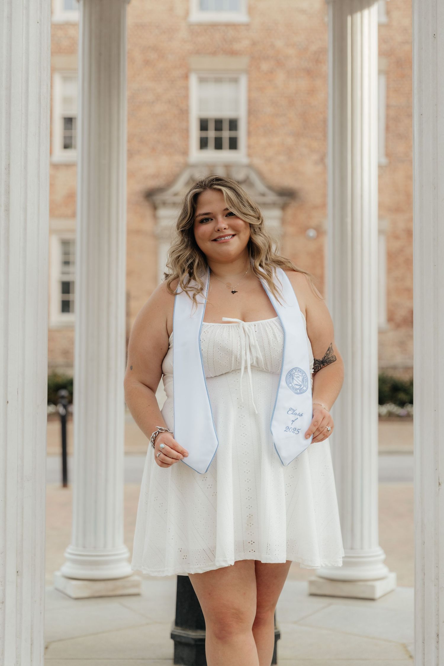 Smiling graduate in white dress holds UNC Chapel Hill graduation stole between white columns on campus.