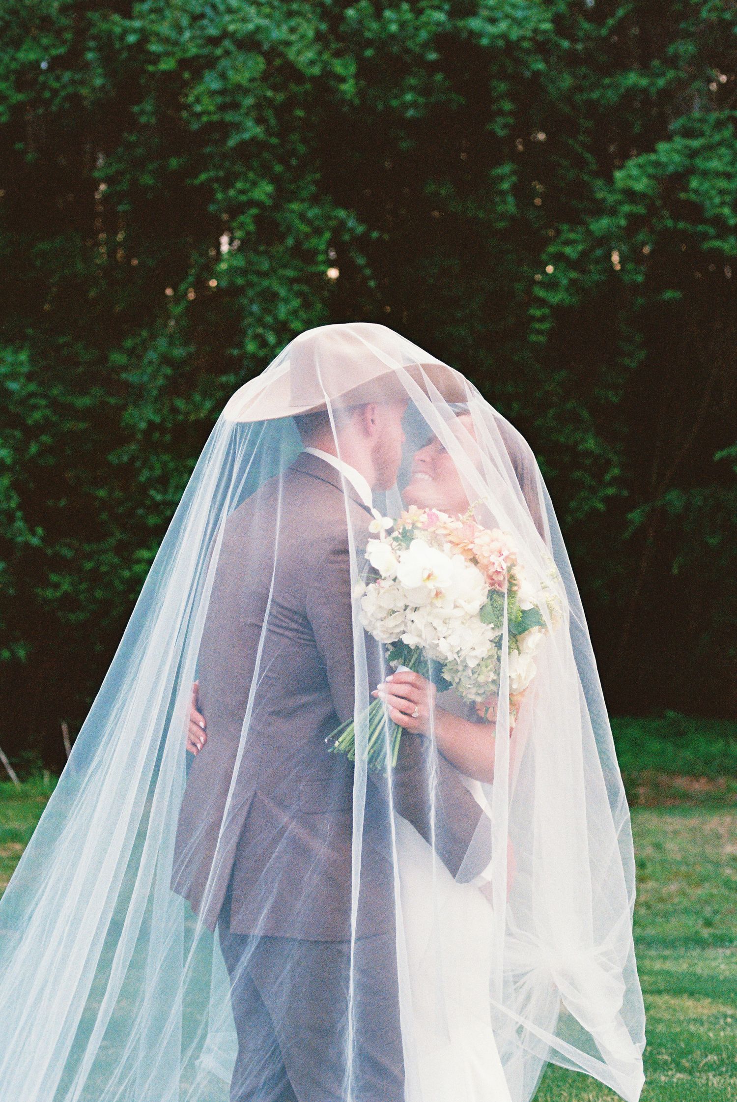 Bride and groom kissing under a flowing white veil, bride holding a white floral bouquet outdoors.