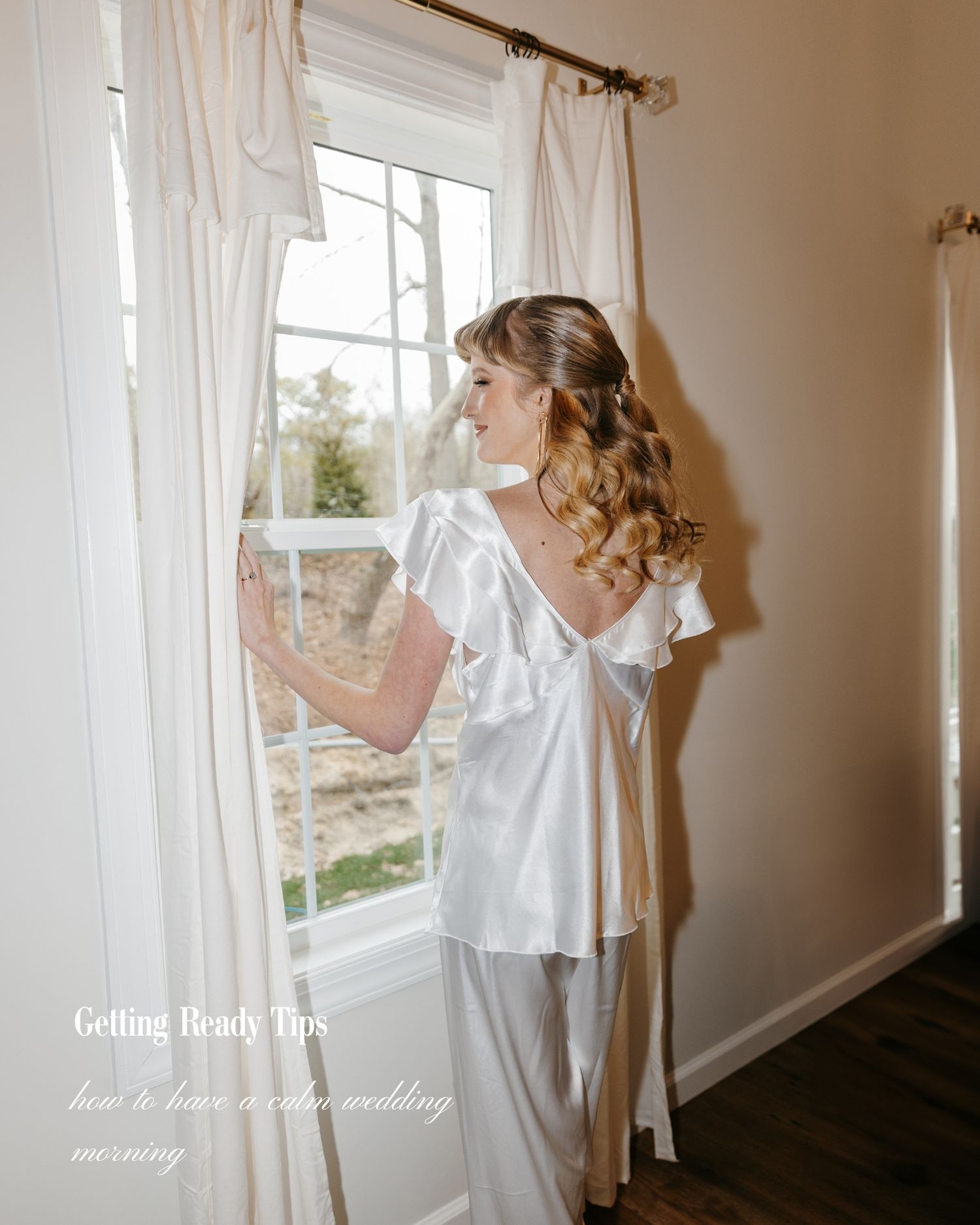 Bride in white satin ruffled pajamas stands by a curtained window on her wedding morning.