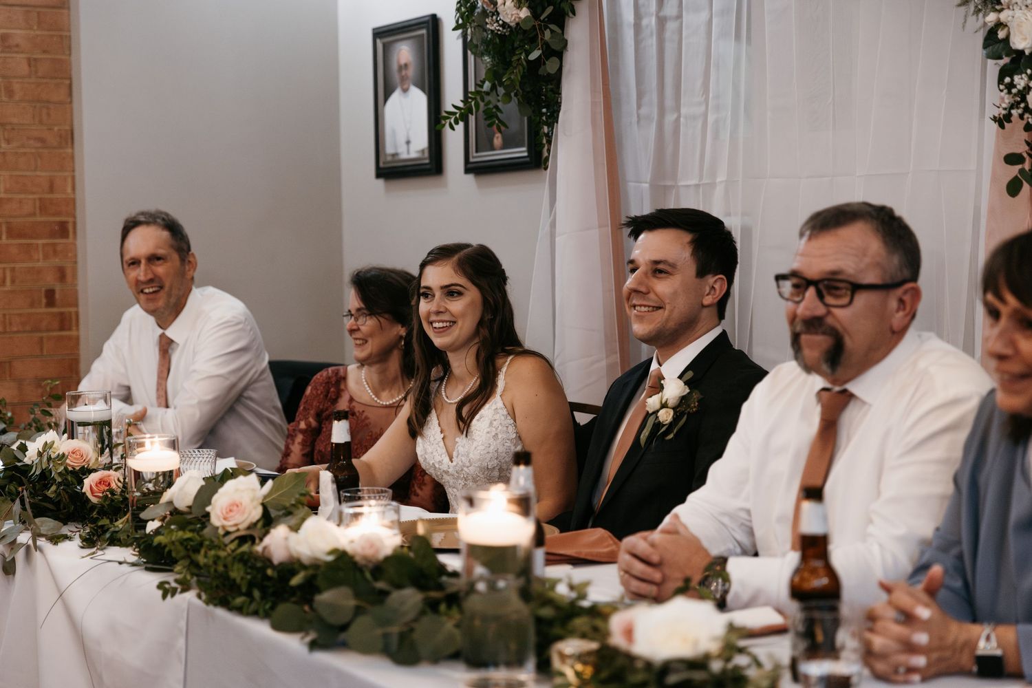 Wedding reception head table setup with floral centerpieces and candlelight against brick walls.
