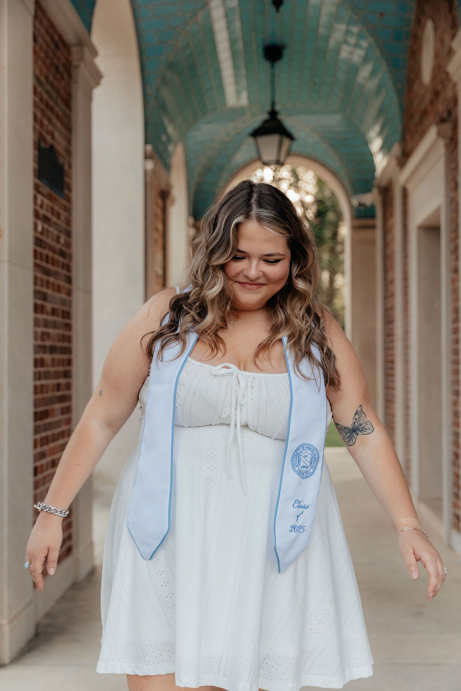 A young woman in a white dress and light blue graduation stole smiles while walking under a brick archway.