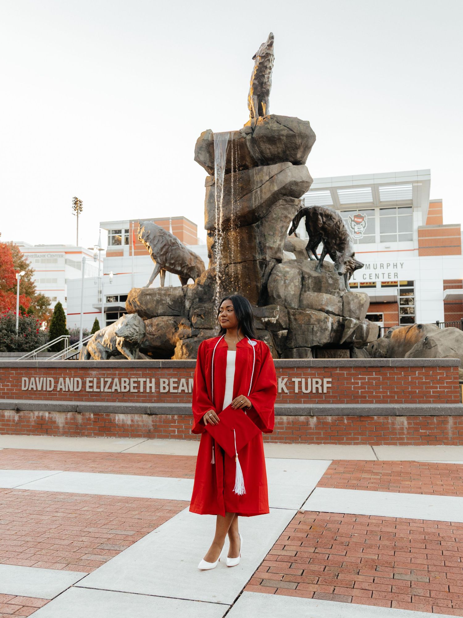 Graduate in red cap and gown poses before a bronze wolf fountain at a university campus plaza.