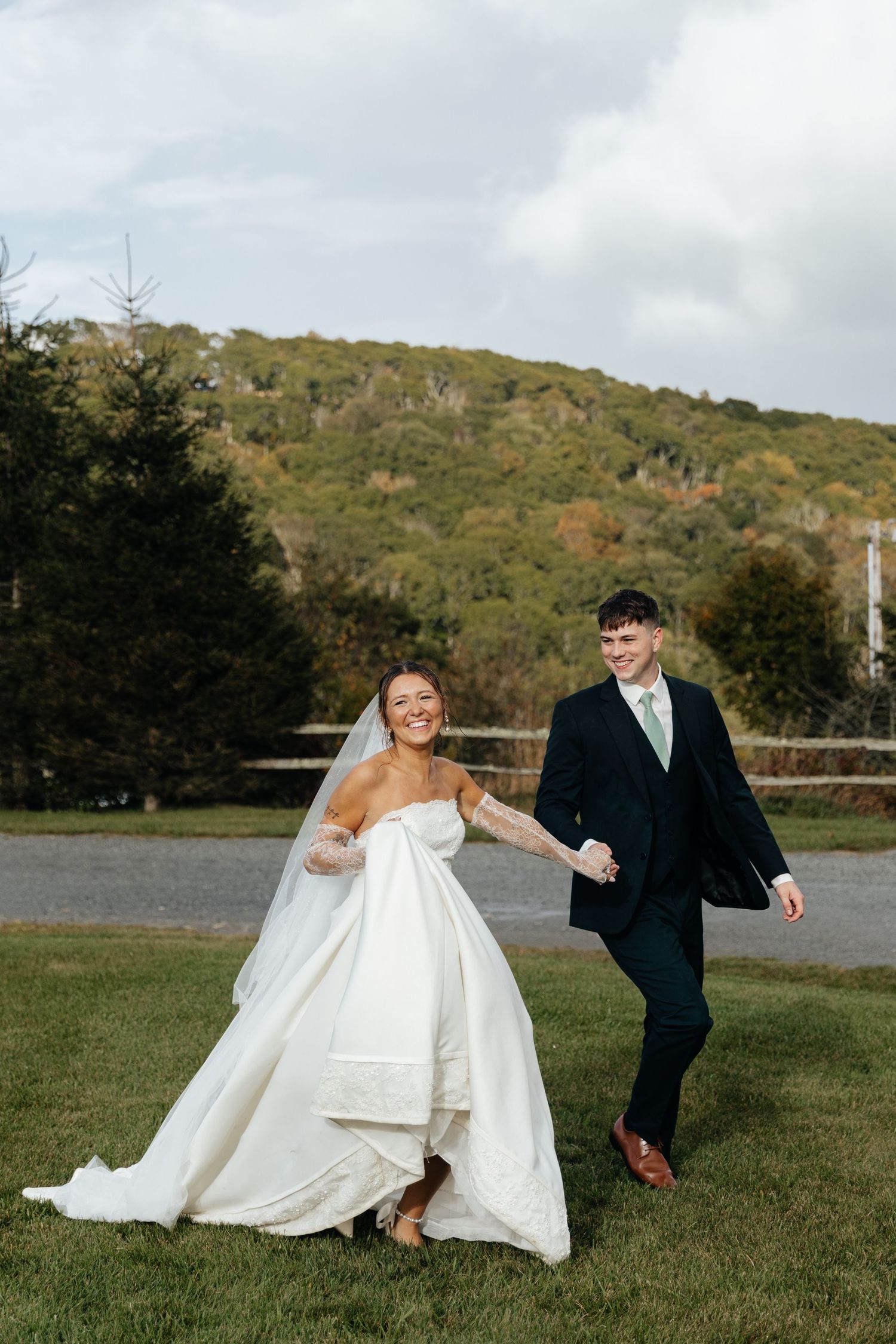 A joyful bride in a white gown and groom in a dark suit walk hand in hand on green grass with a wooded hill backdrop.