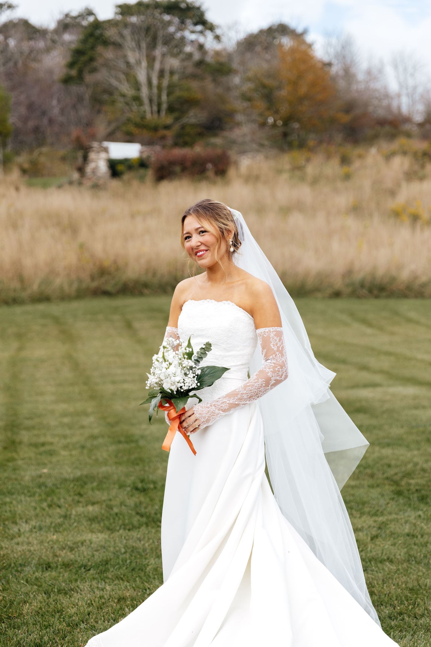 Smiling bride in strapless white gown with lace sleeves, veil, and bouquet standing on green lawn with autumn trees.