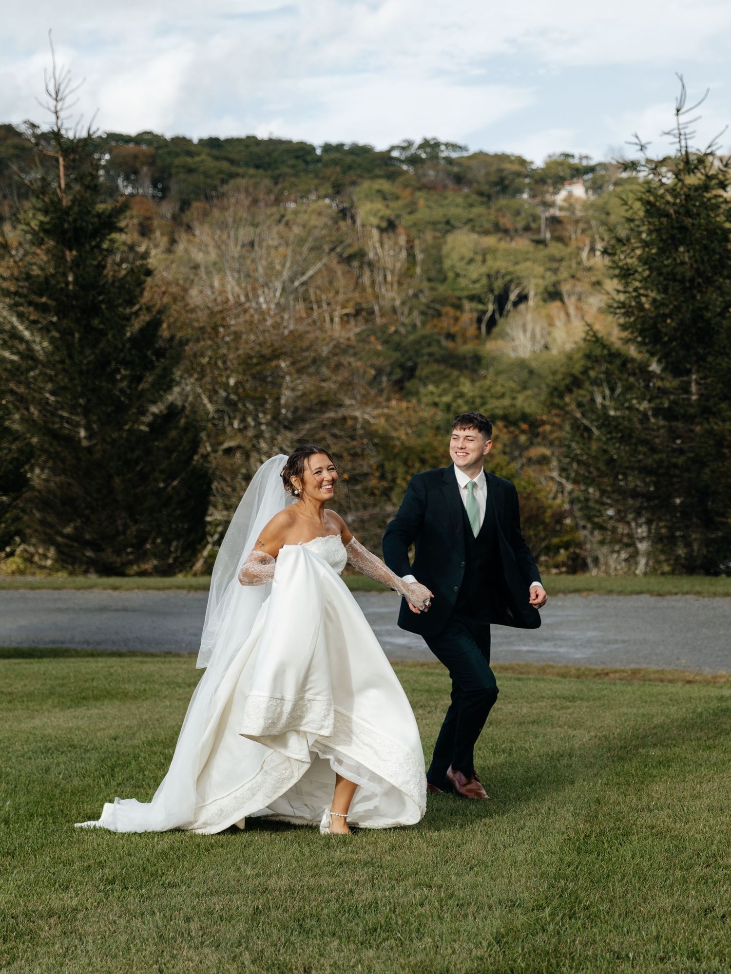 A joyful bride and groom walk hand-in-hand on green grass with autumn trees and hills in the background.
