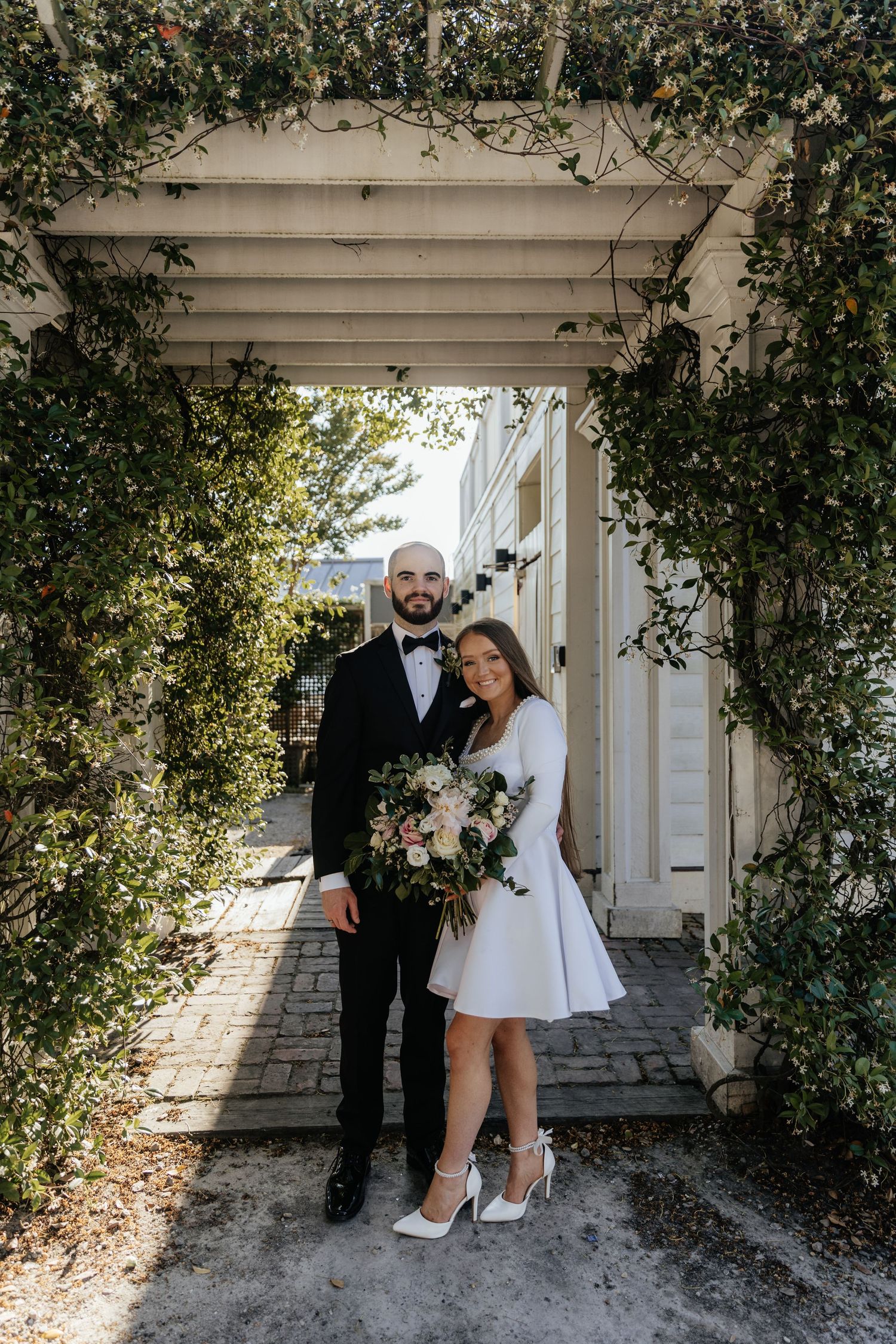 A couple poses under a vine-covered archway holding a lush white and pink floral bouquet.