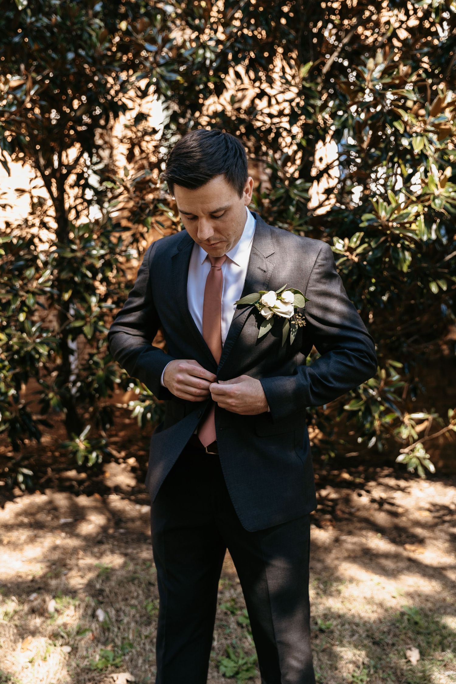 Groom in dark suit with coral tie standing among trees and foliage.