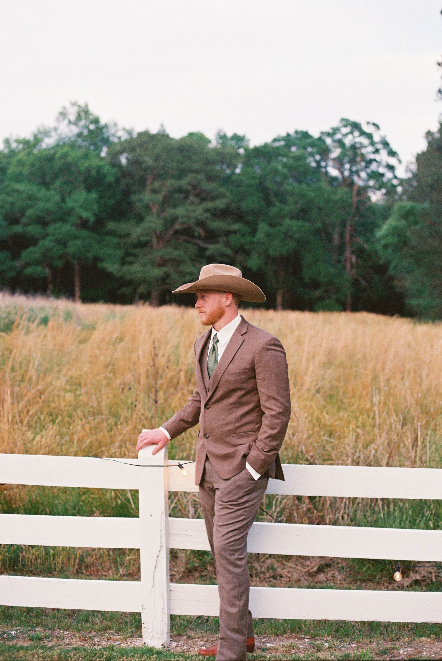 A man in a brown suit and cowboy hat leans on a white fence in a golden field.
