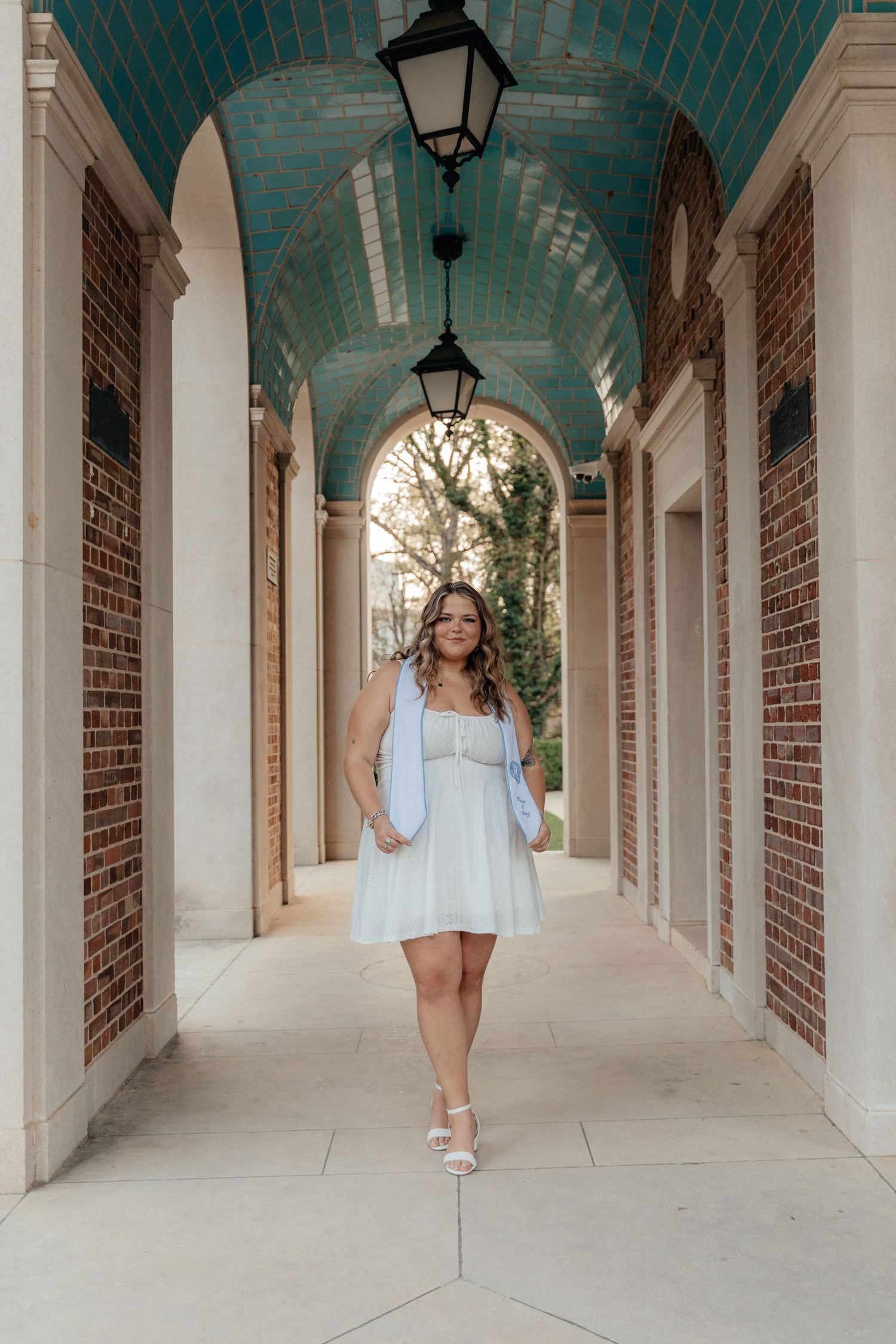 Young woman in light blue mini dress walks elegant brick archway with ornate teal ceiling and hanging lanterns.