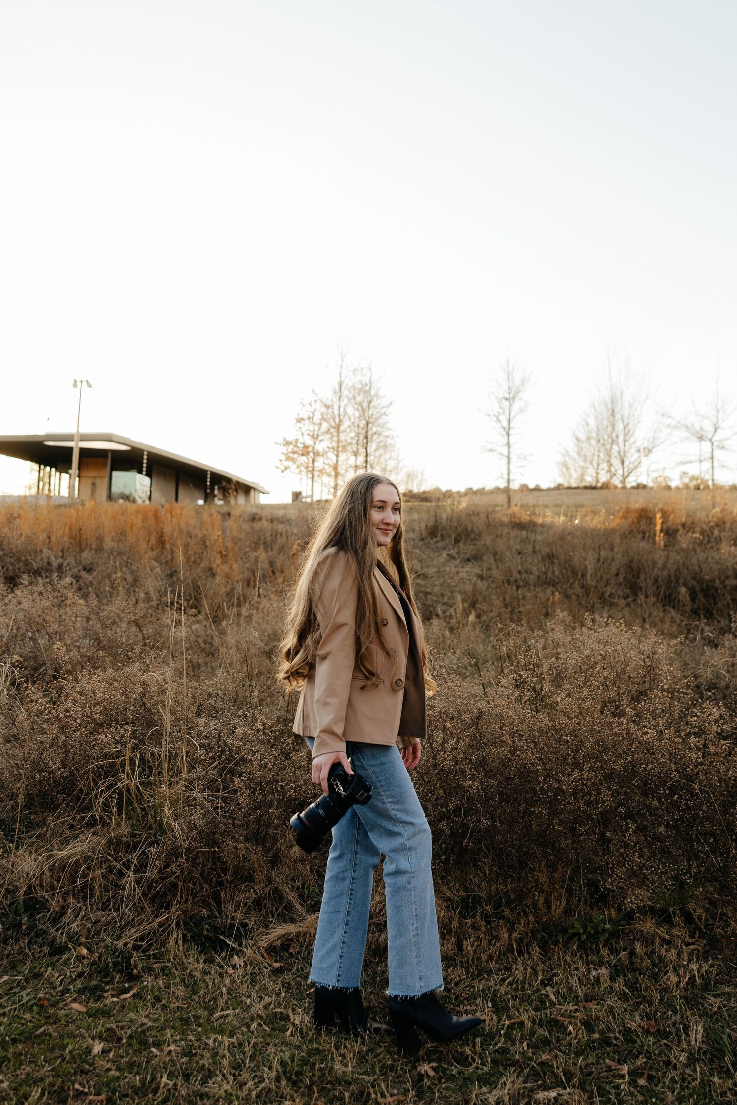 Person in tan coat and jeans stands in autumn field during golden hour.