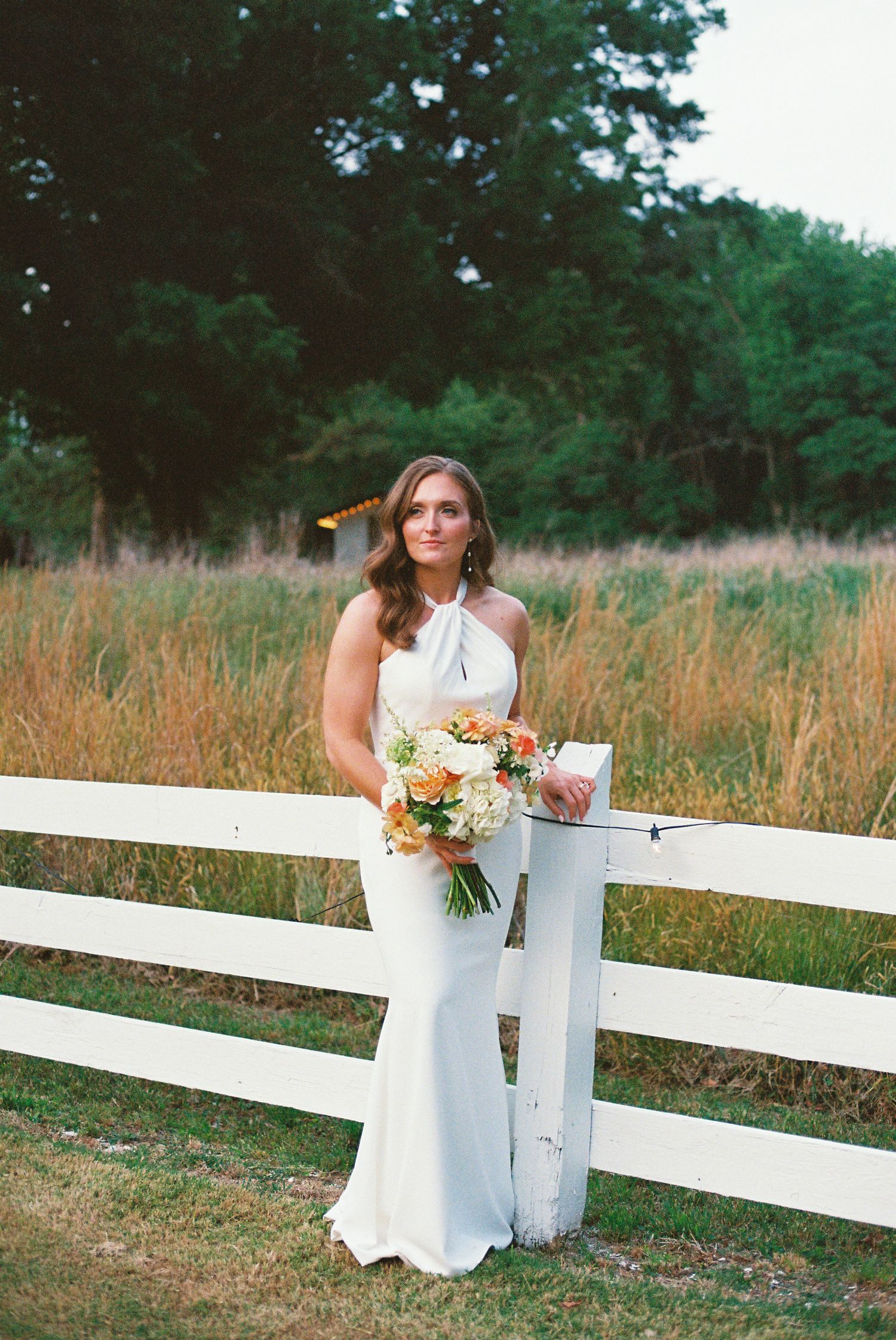 Bride in white halter gown holding orange and white bouquet by white fence in rustic outdoor setting.