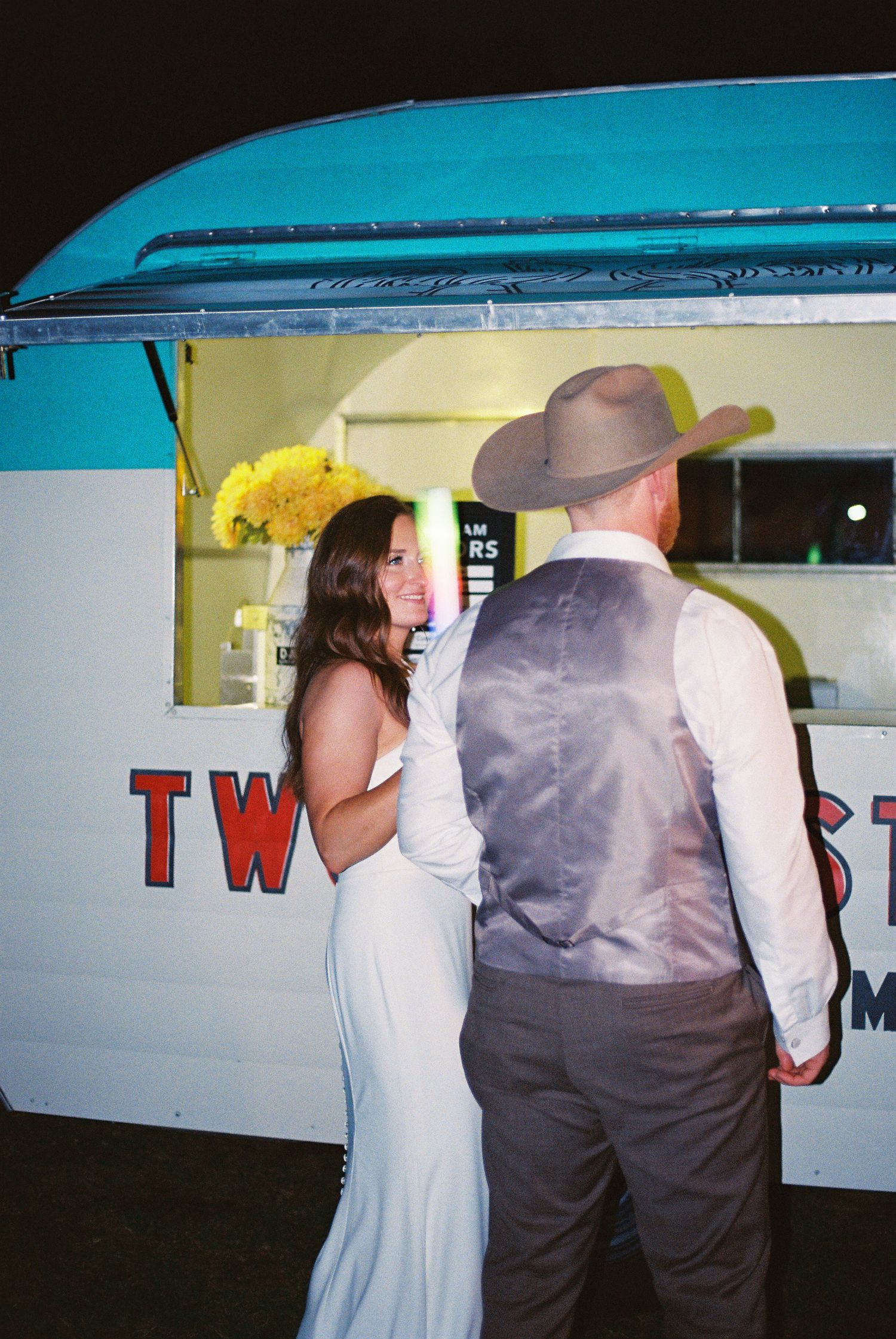 Couple in wedding attire at a food truck, groom wearing cowboy hat and vest, bride in elegant white gown.