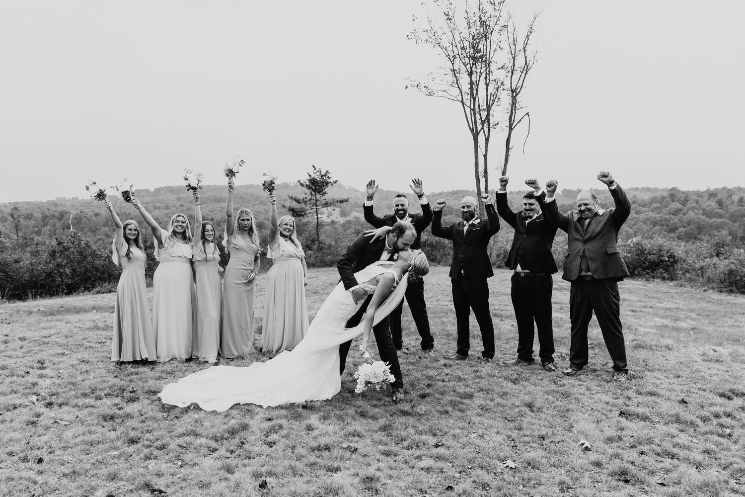 Wedding party poses together in black and white on grassy hillside while bride and groom share a kiss.