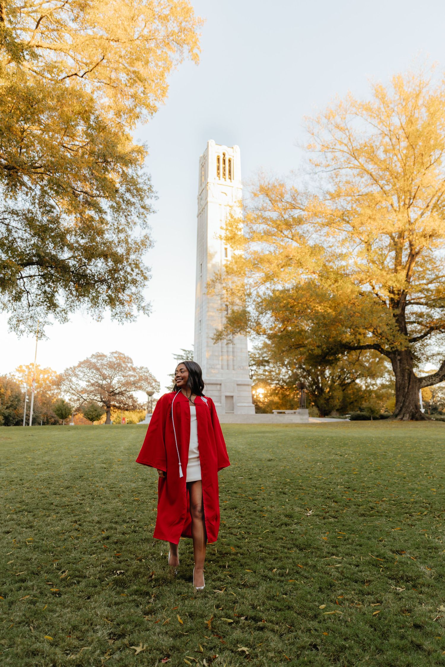 Graduate in red cap and gown walks across green campus lawn with tall bell tower in background.