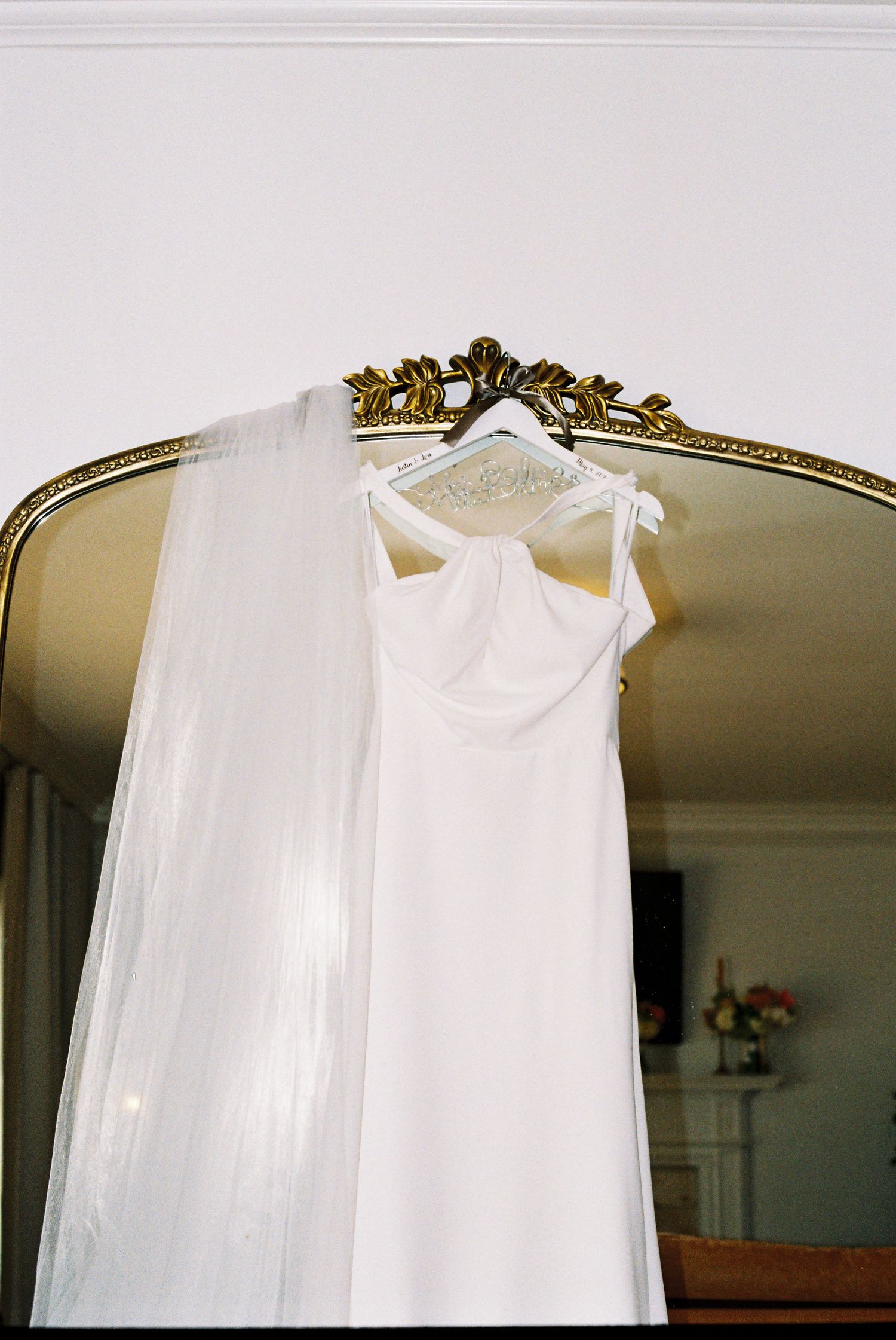 White wedding dress and veil hanging on ornate gold mirror frame in elegant bridal suite.