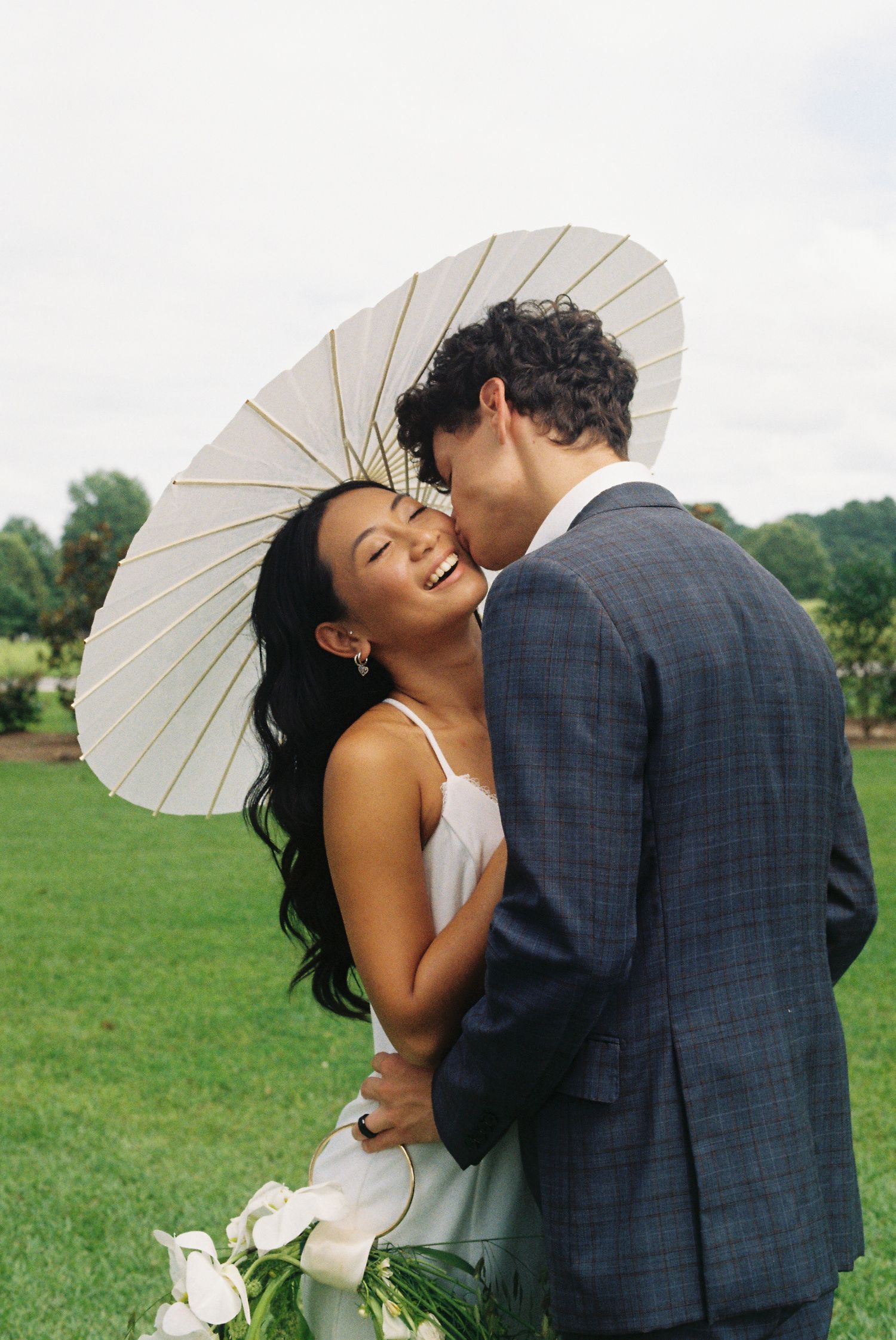 Wedding couple shares romantic moments with white parasol on green lawn.