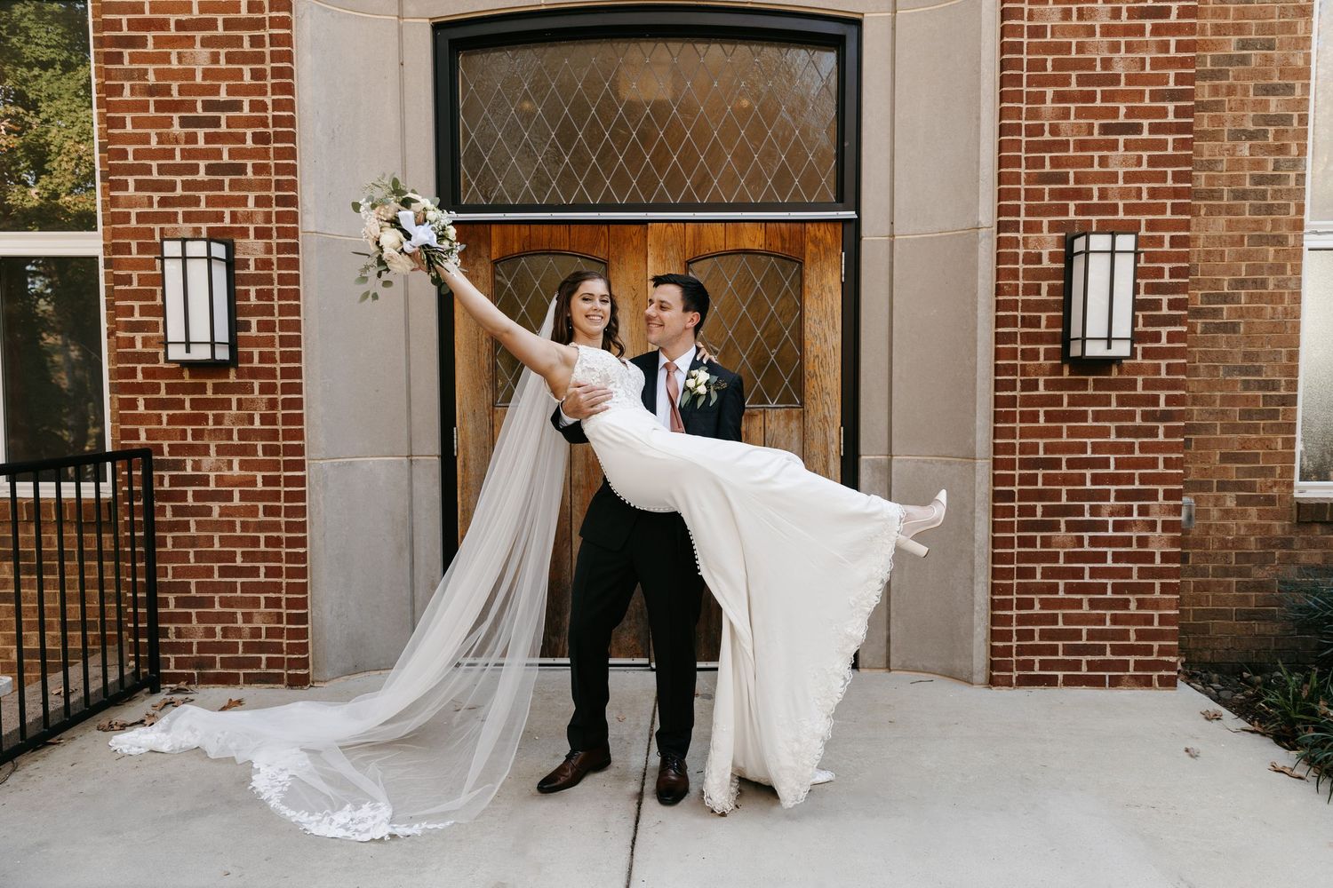 Dramatic wedding pose with flowing veil in front of brick building entrance.