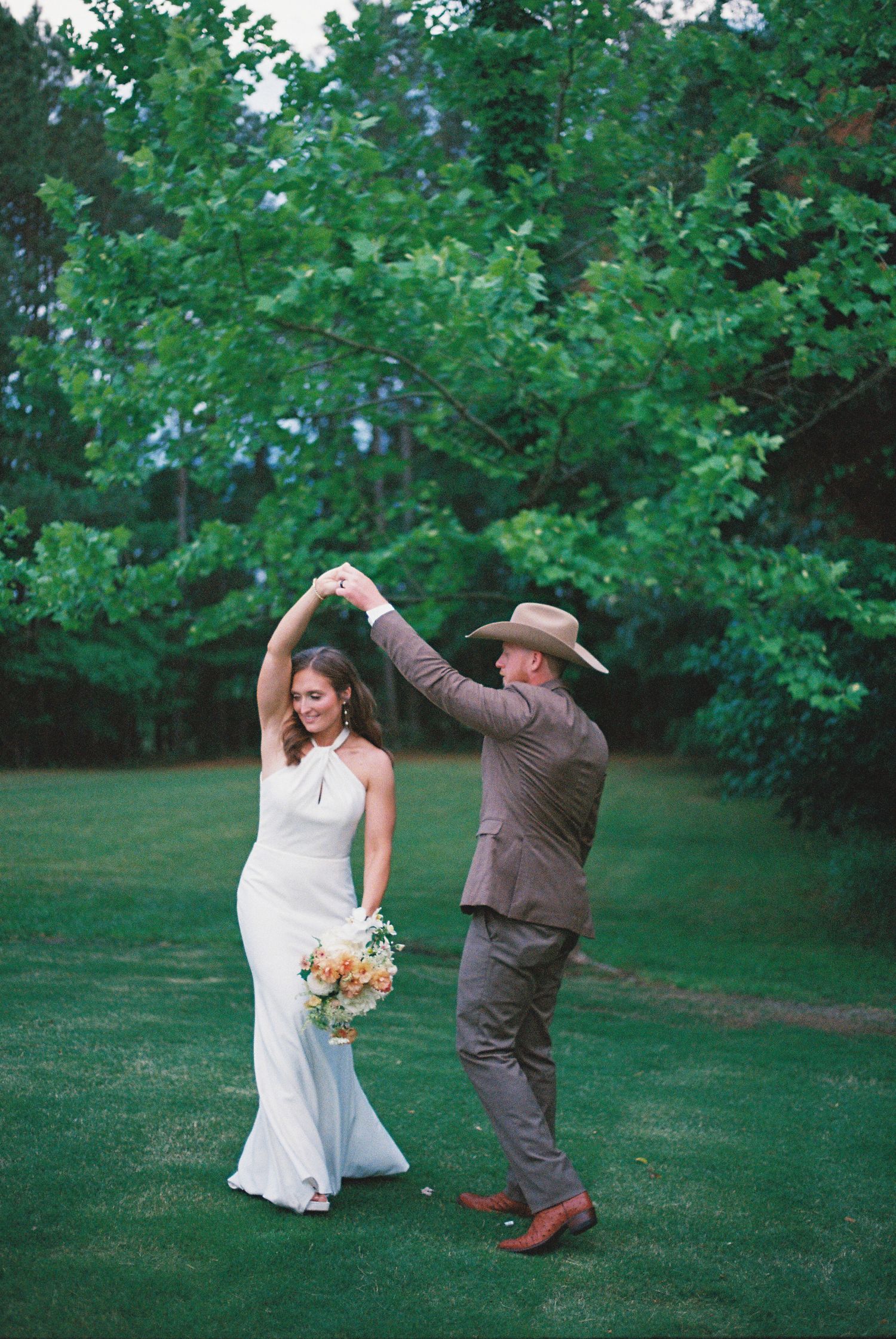 Bride and groom dancing outdoors on green lawn, groom in tan cowboy hat and suit, bride in white halter gown holding bouquet.