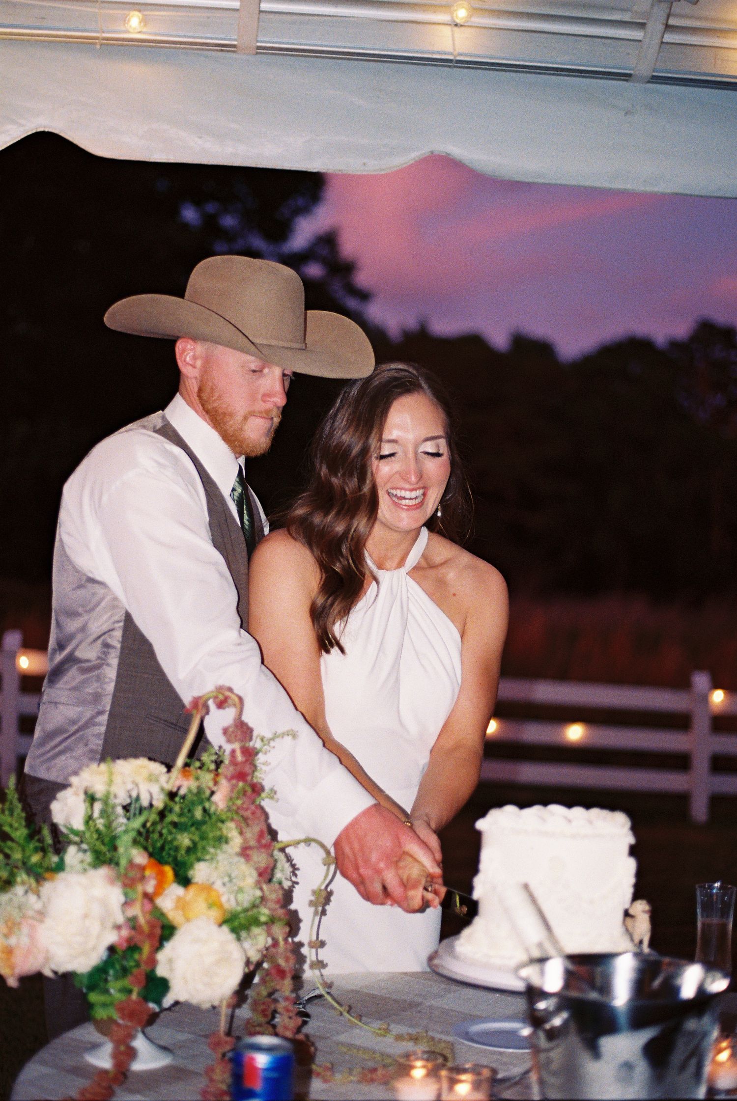 Bride and groom cutting their white wedding cake outdoors at sunset, groom in cowboy hat and gray vest.