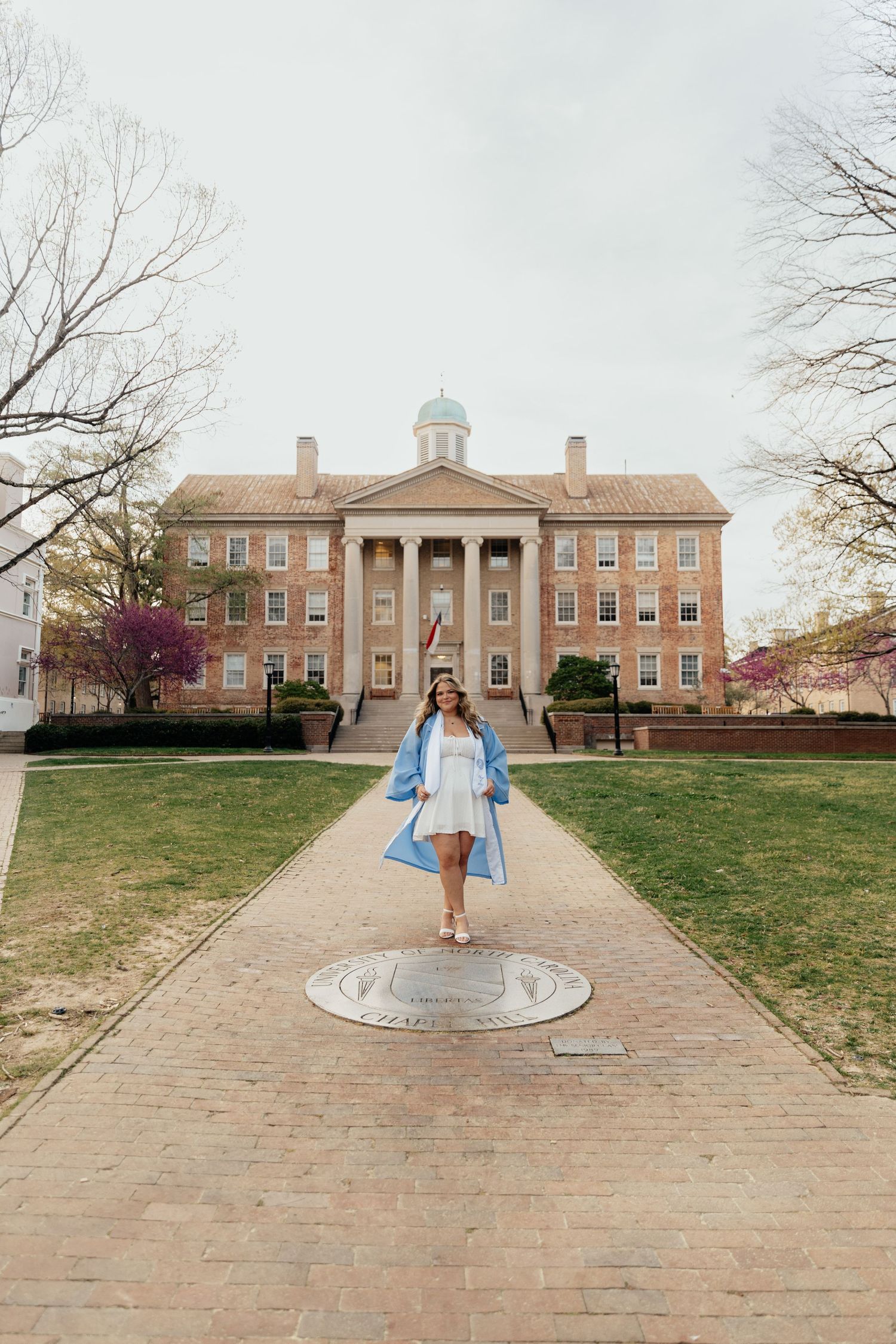 Graduate poses in white dress and blue robe at UNC Chapel Hill seal with historic South Building behind her.