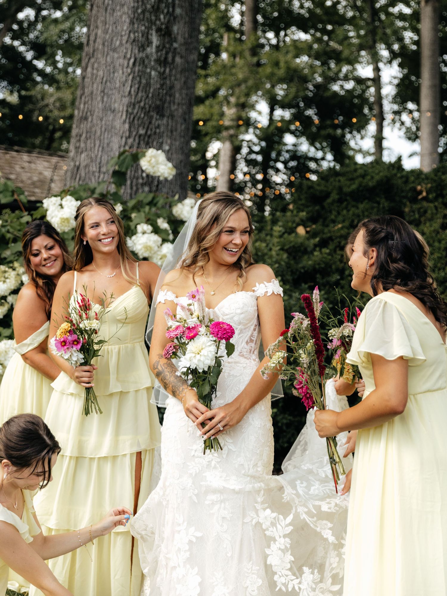 Bridal party shares joyful moment while helping with bride's dress in garden setting.