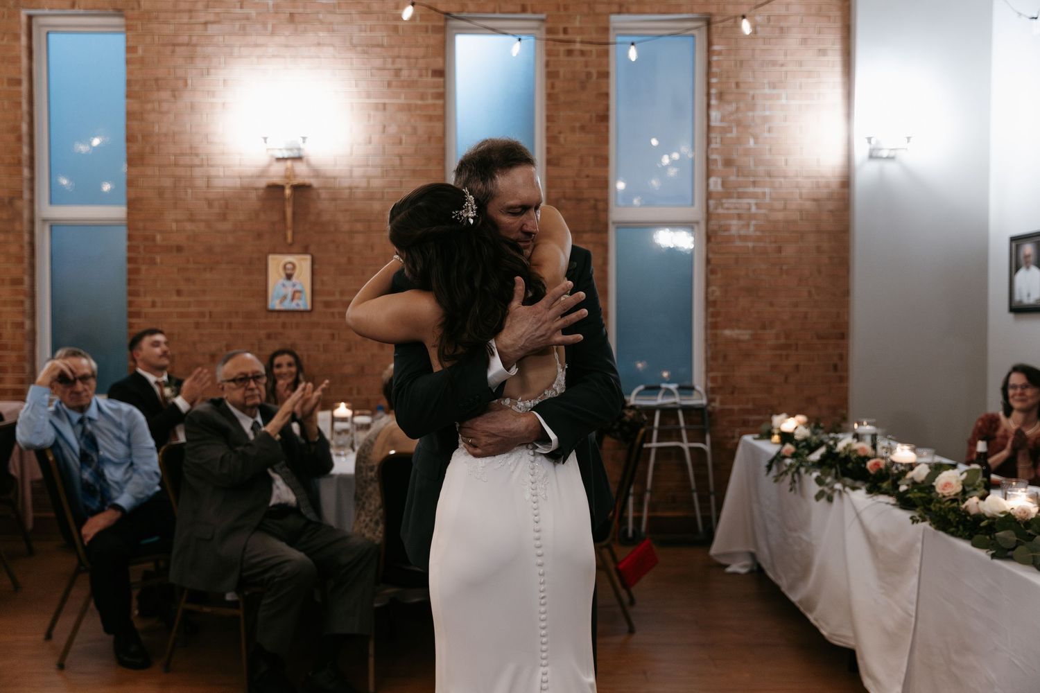 Bride and her father dancing and celebrating in a rustic brick-walled venue with string lights overhead.