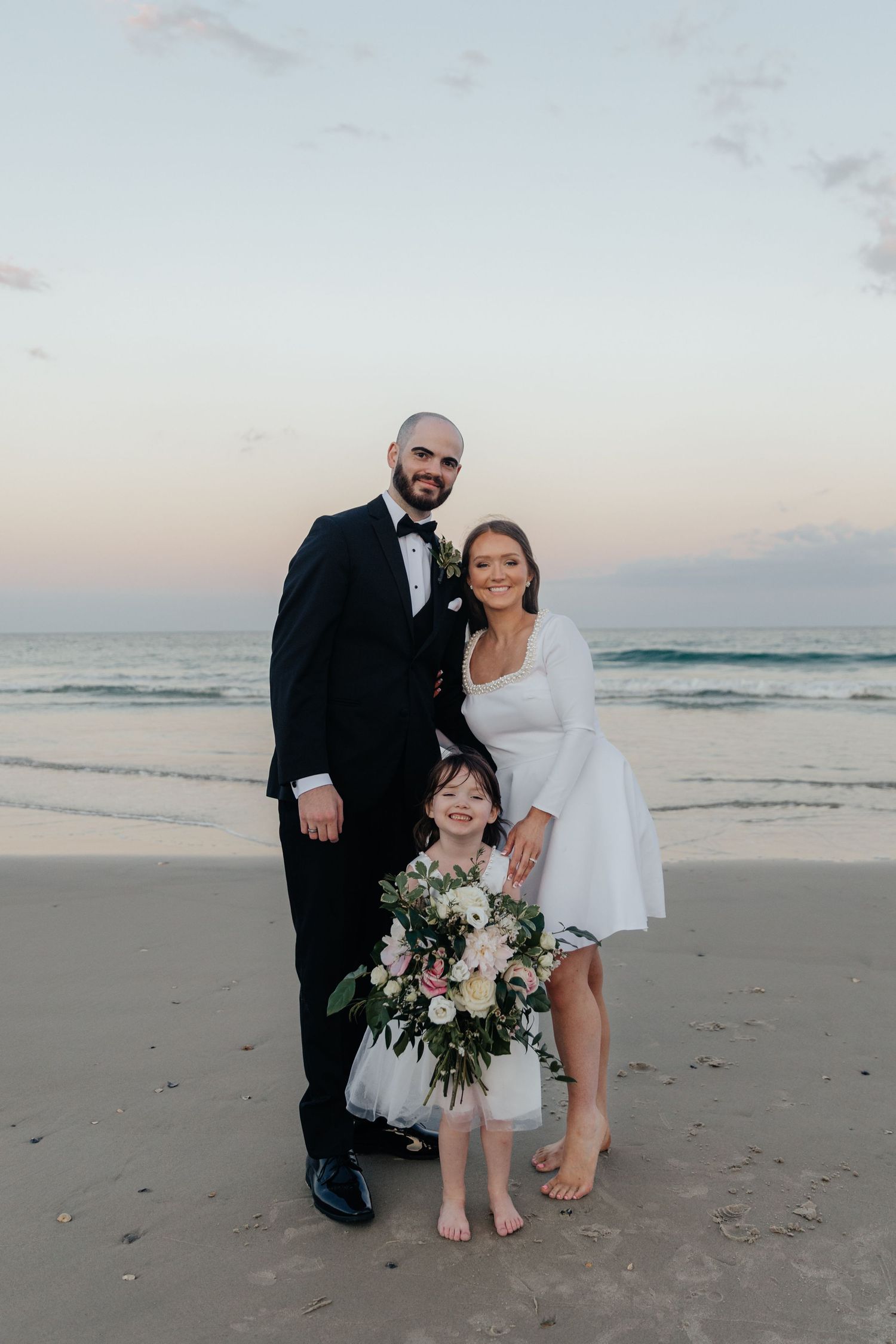 A wedding family portrait on the beach at sunset with a beautiful floral bouquet.
