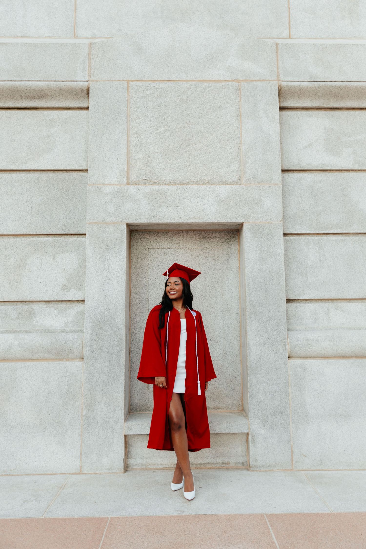 A graduate poses in a red cap and gown against a white stone wall.