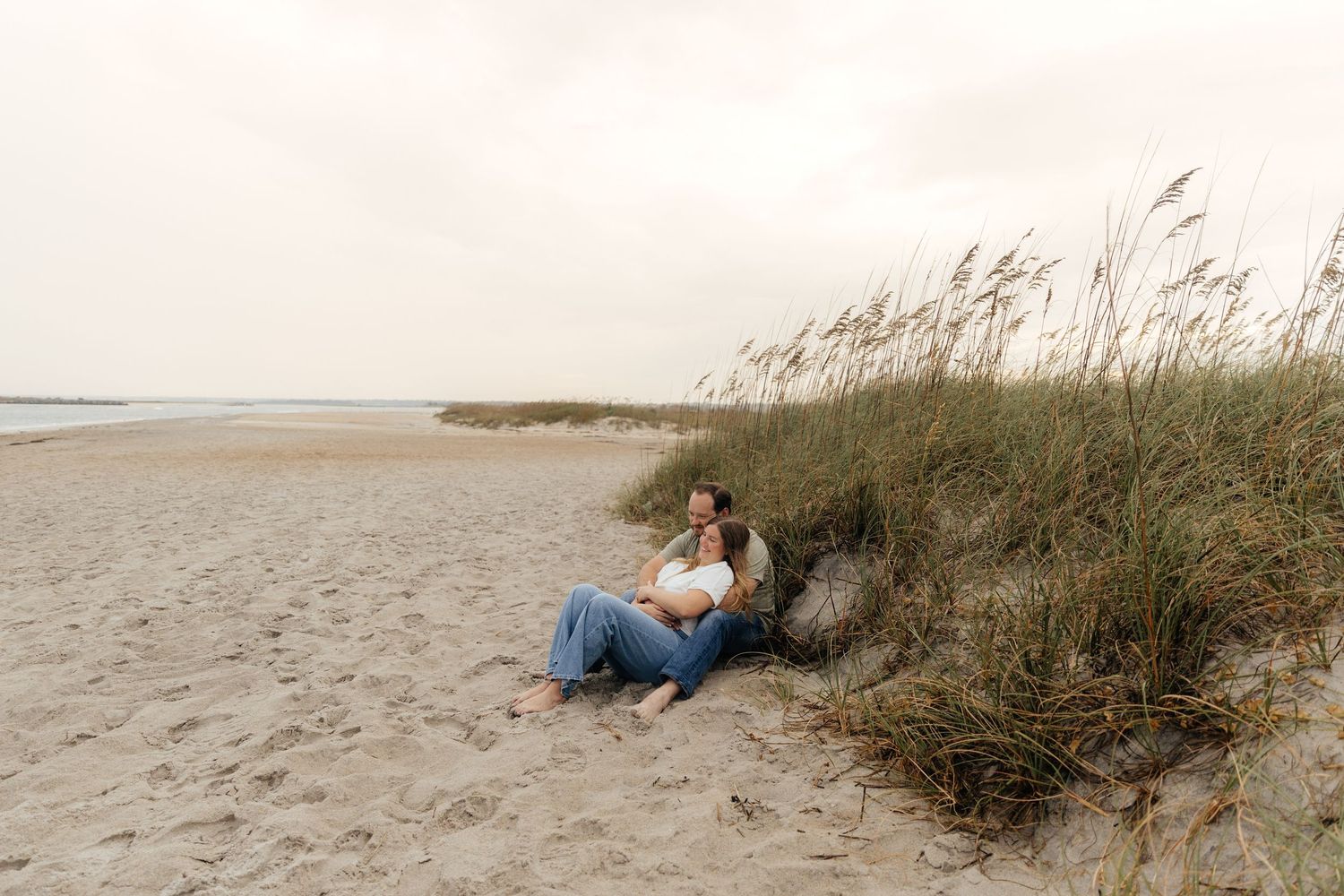 Couple relaxes on sandy beach near grass during coastal photoshoot.