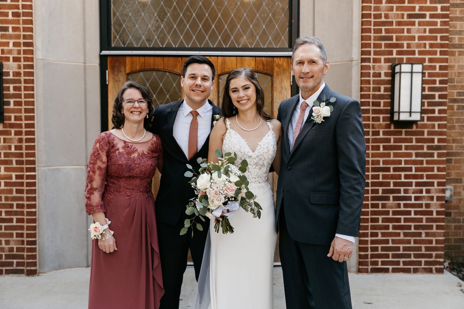 Wedding party poses outside a brick church entrance with floral bouquets and burgundy bridesmaid dresses.