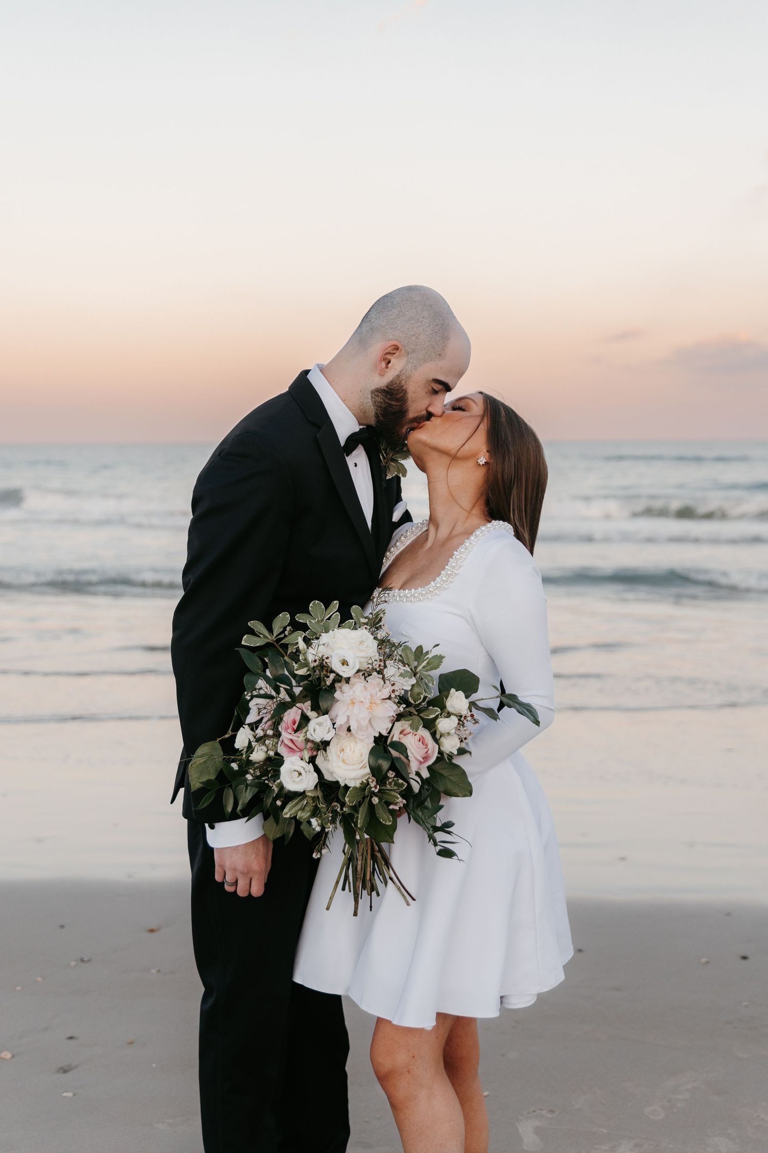 A romantic beach wedding photo of a couple sharing a kiss at sunset with white floral bouquet.