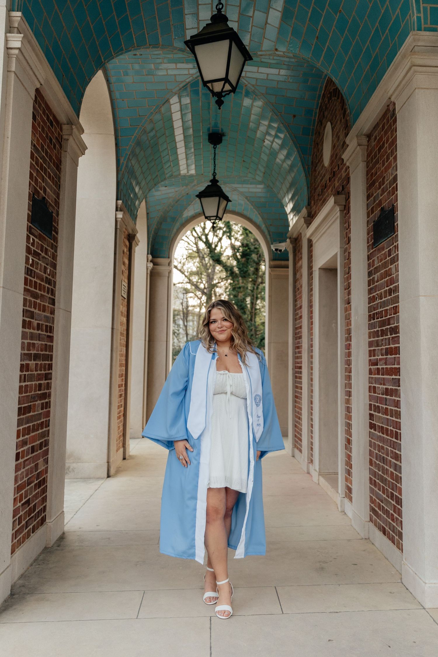 Young woman in light blue graduation gown and white dress walking through brick archway corridor.
