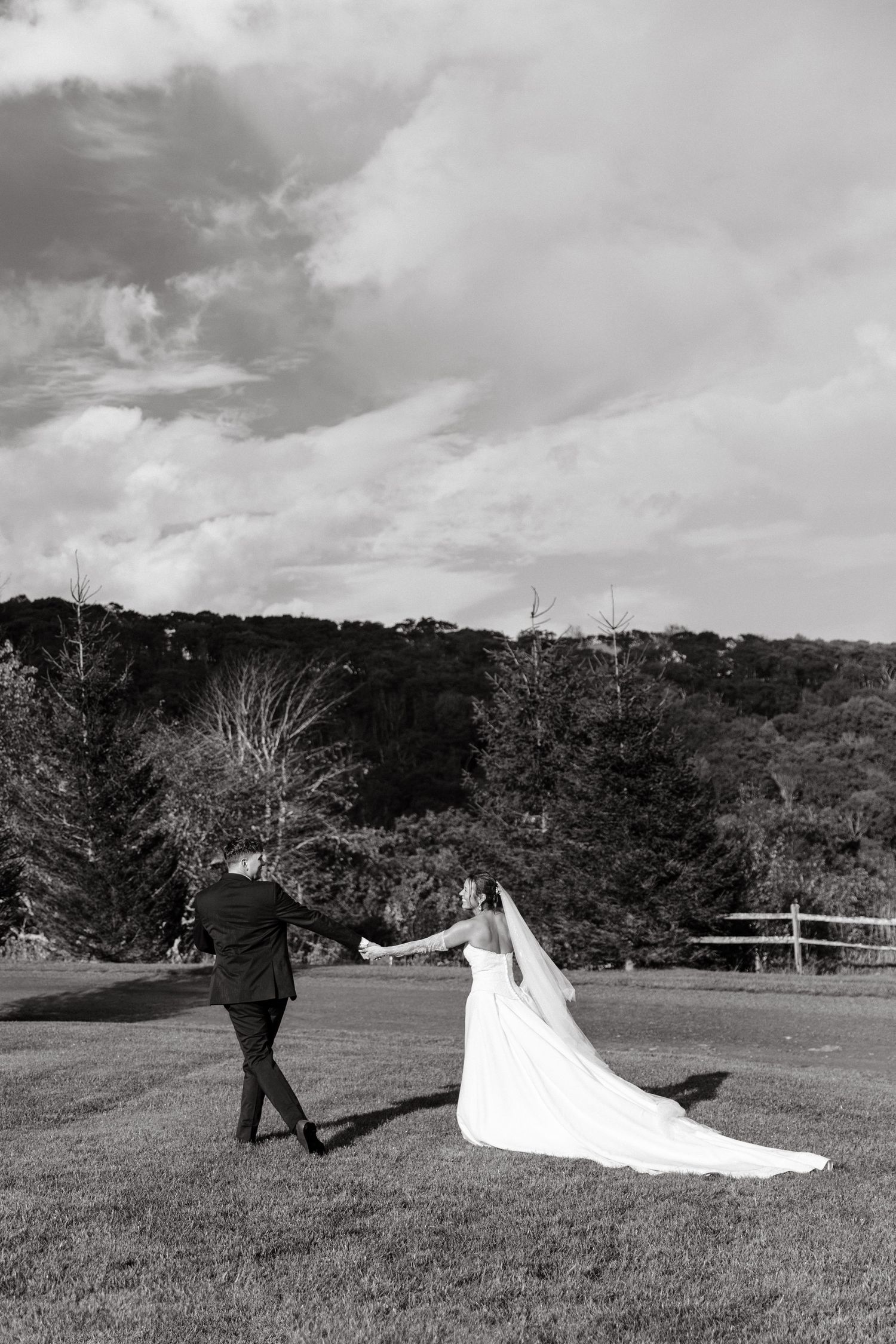 Black and white photo of bride and groom holding hands walking across a lush green lawn with trees in the background.