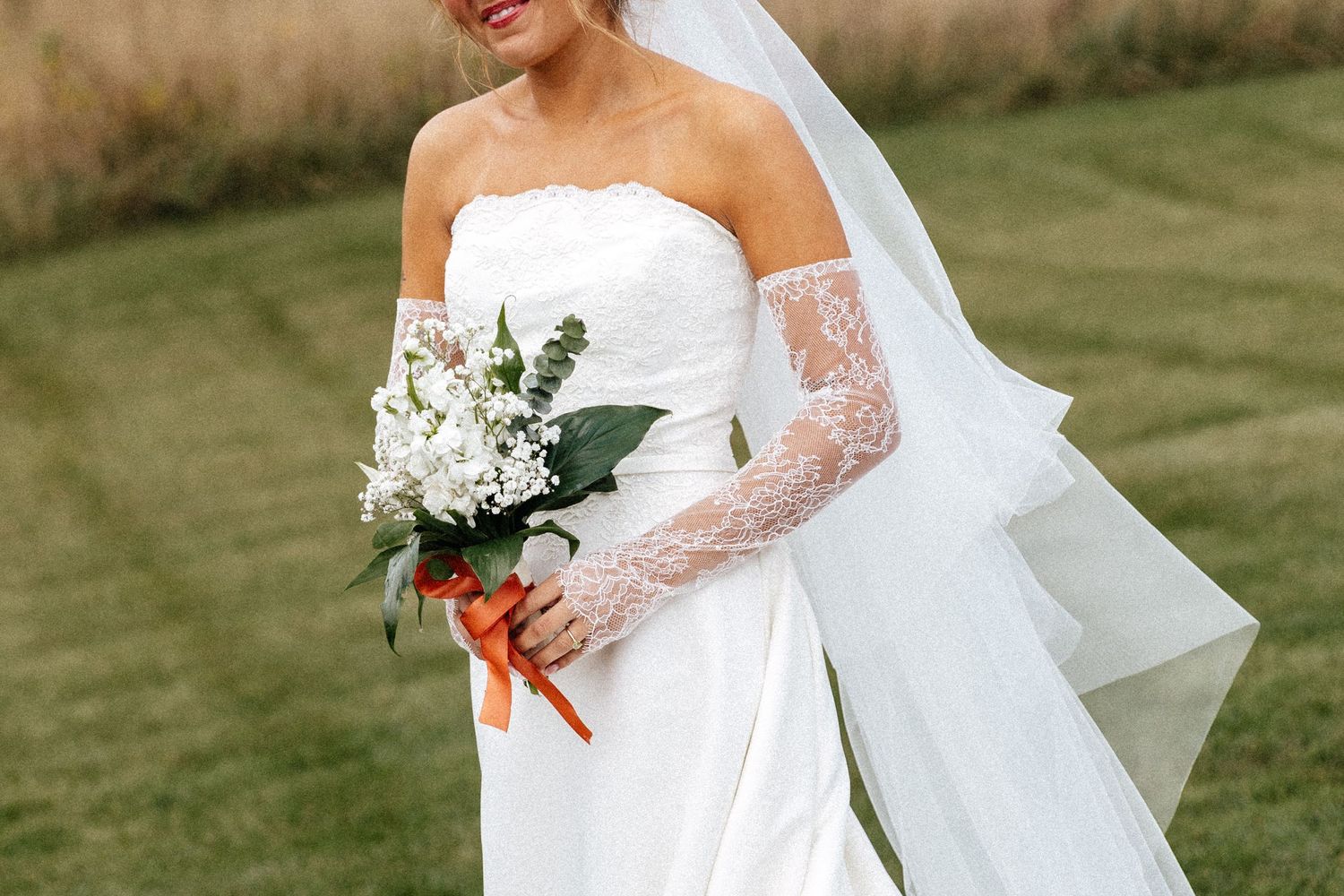 Bride in strapless white gown with lace gloves and veil holds white floral bouquet with orange ribbon outdoors.