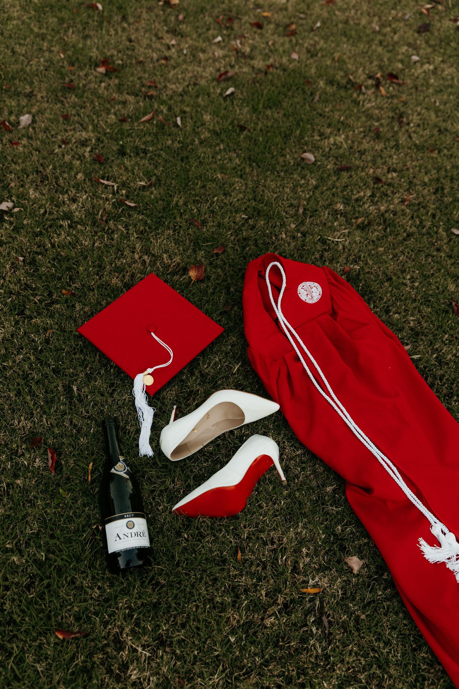 Red graduation cap, gown, white heels, and Andre champagne bottle on green grass for a celebratory grad photoshoot.