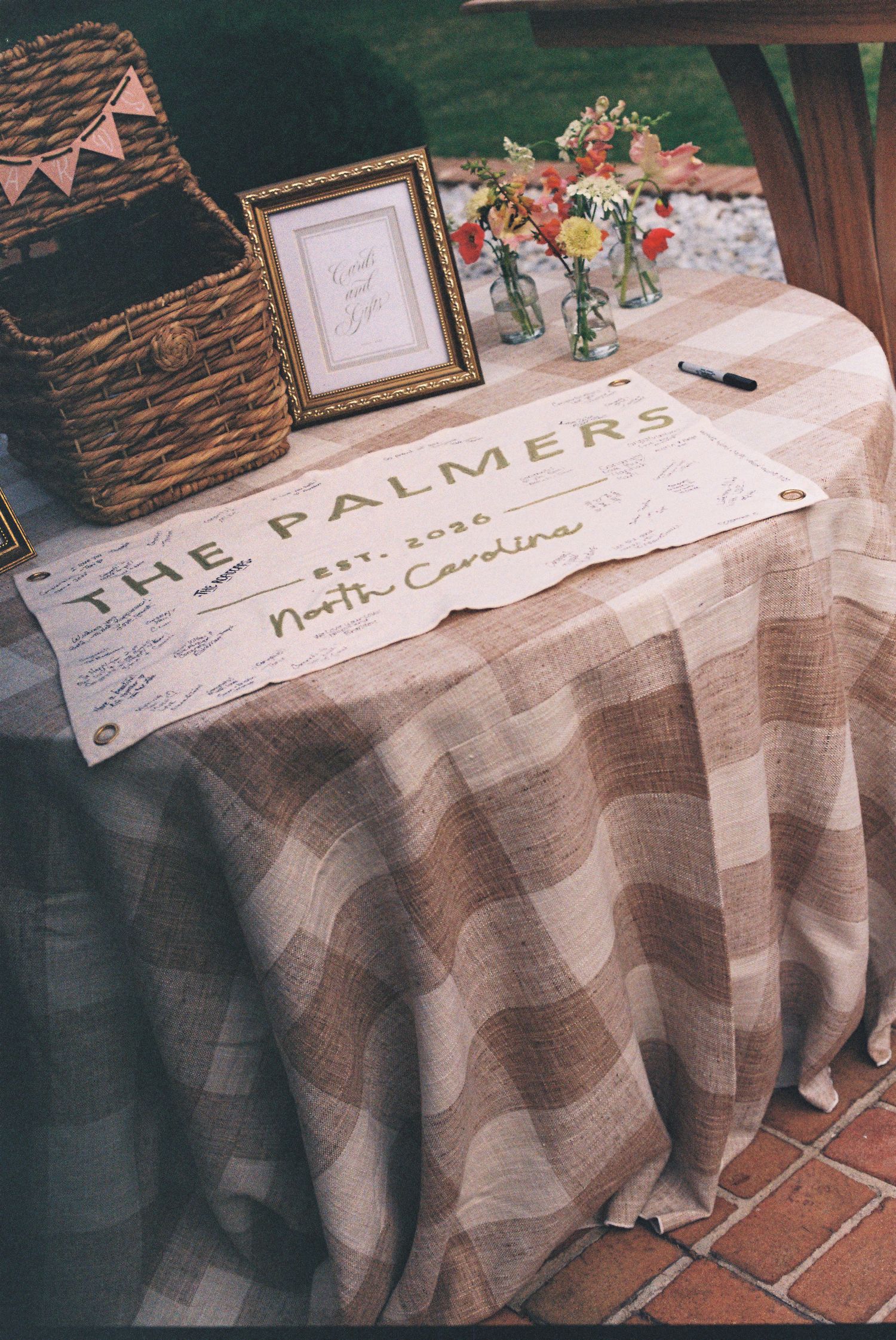 Rustic wedding guest sign-in table with The Palmers Est. 2020 North Carolina banner, plaid tablecloth, and floral arrangement.