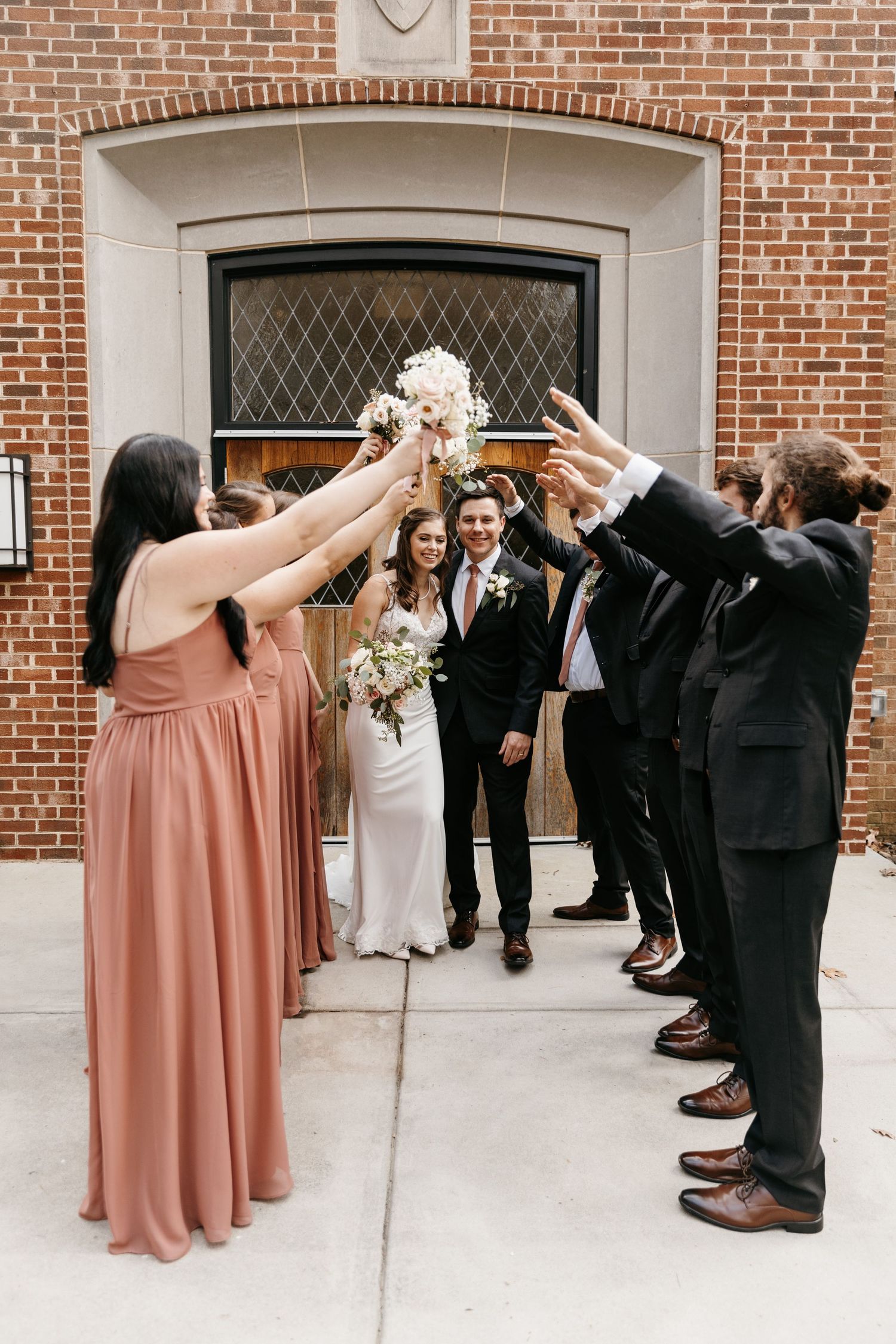 Bridal party forms celebratory arch with raised arms against red brick wall and arched doorway while bride and groom walk through it.