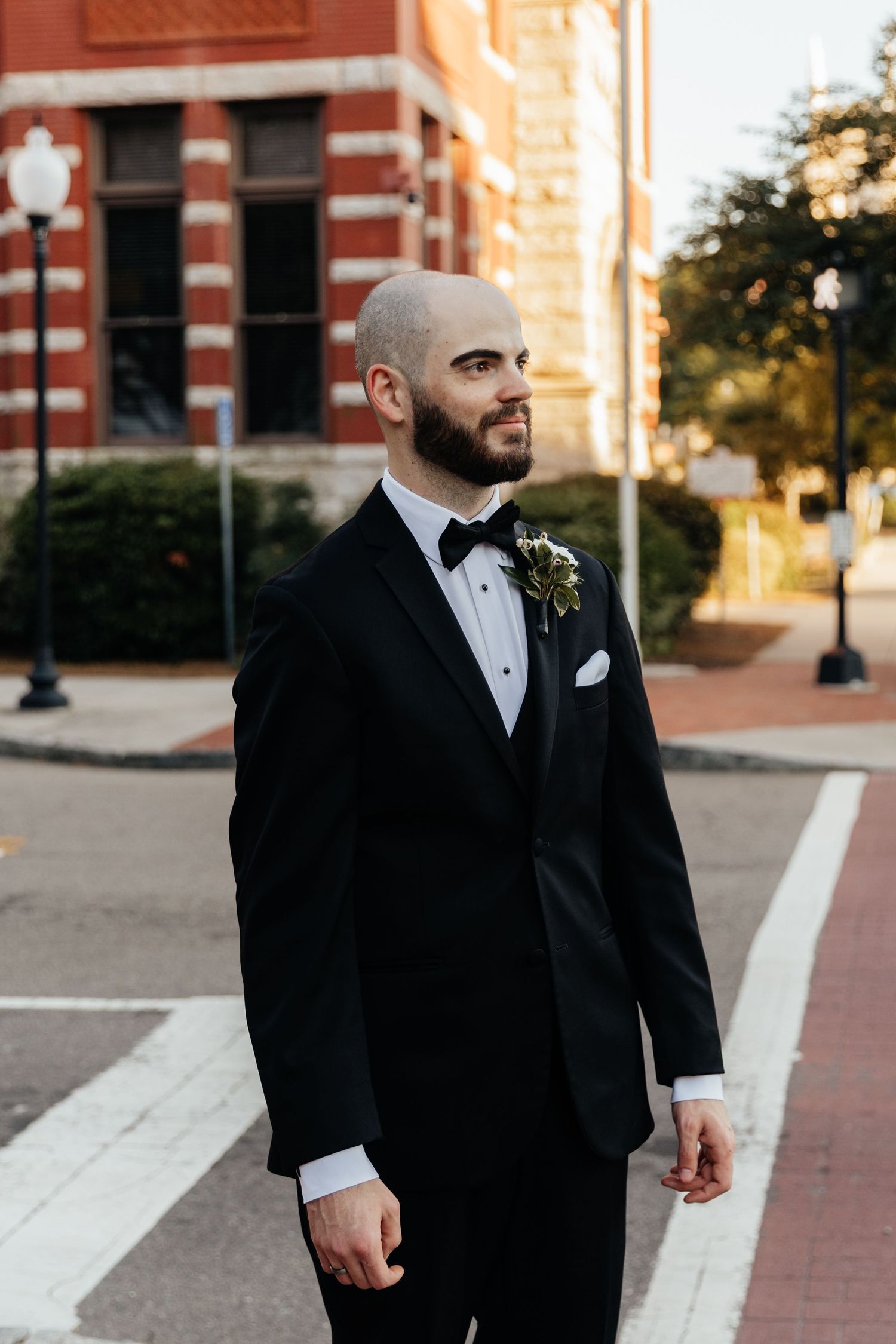 A groom in a classic black tuxedo poses outdoors near brick buildings during a downtown wedding.