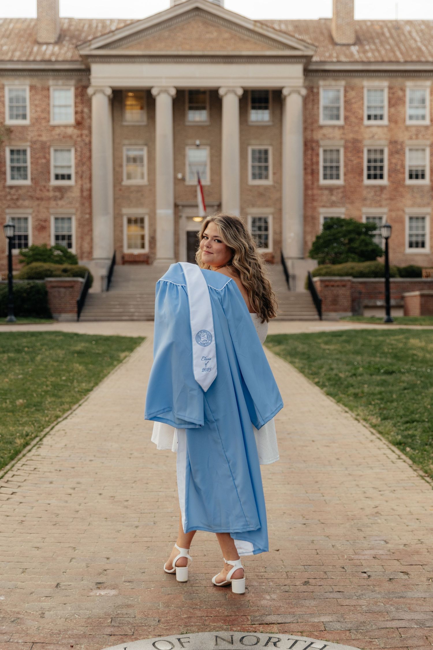 Graduate in light blue gown twirls on campus walkway in front of classic brick university building.