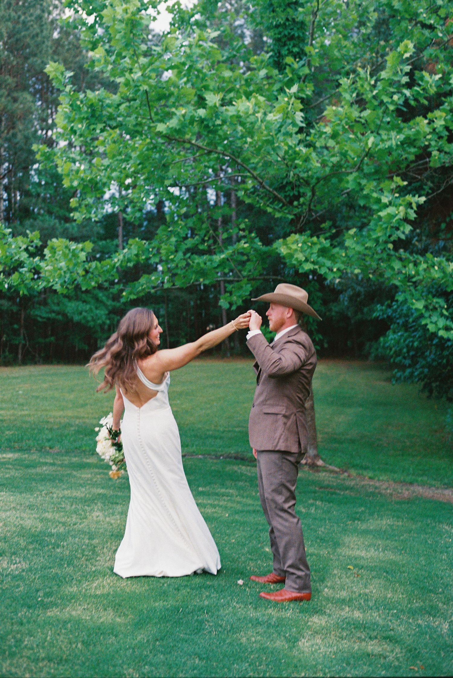 Bride and groom dancing outdoors on green lawn surrounded by lush trees during rustic wedding celebration.