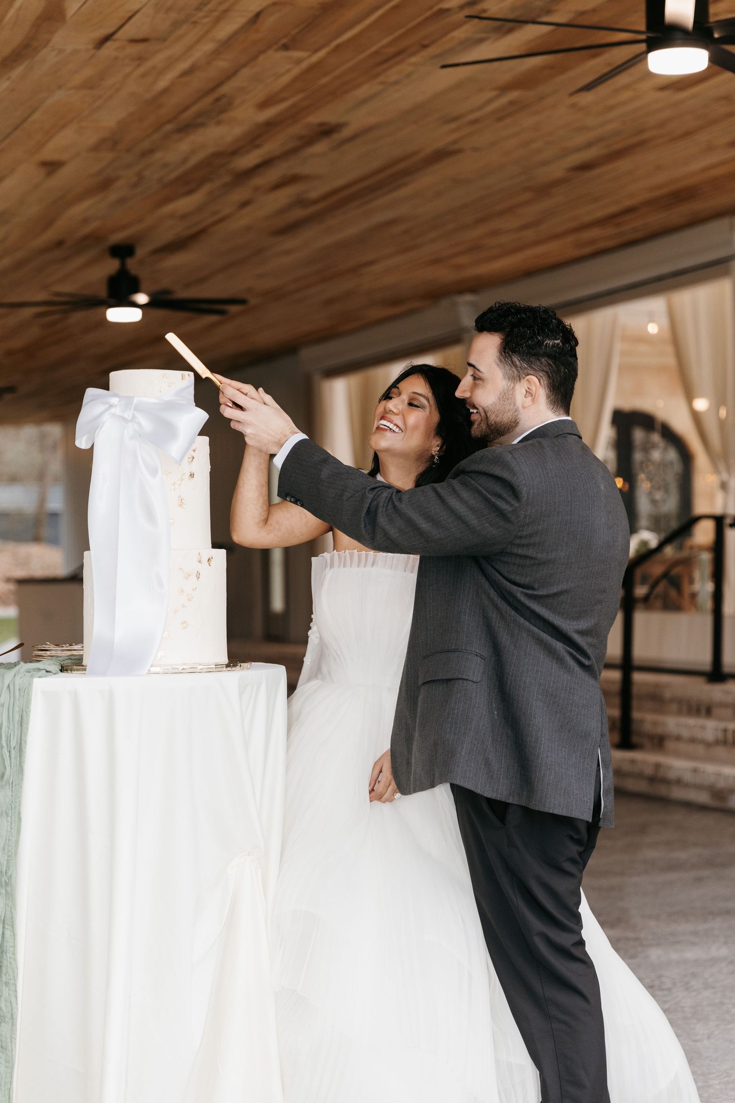 Bride and Groom cutting wedding cake captured under wooden beamed outdoor structure.