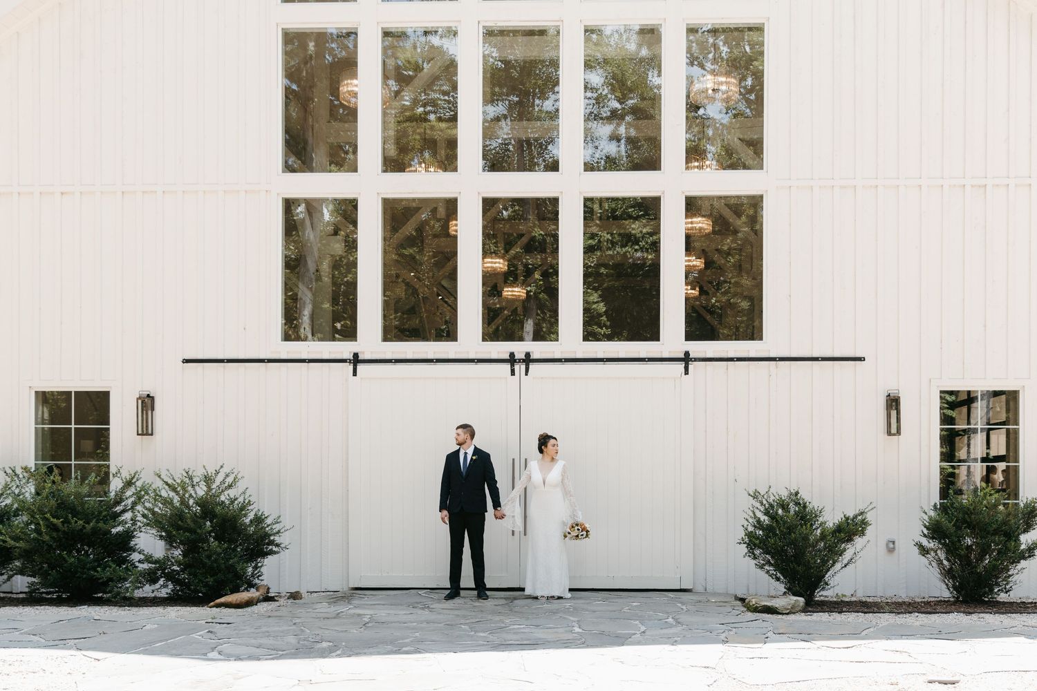 A white venue exterior with tall windows serves as backdrop for a wedding couple portraits.